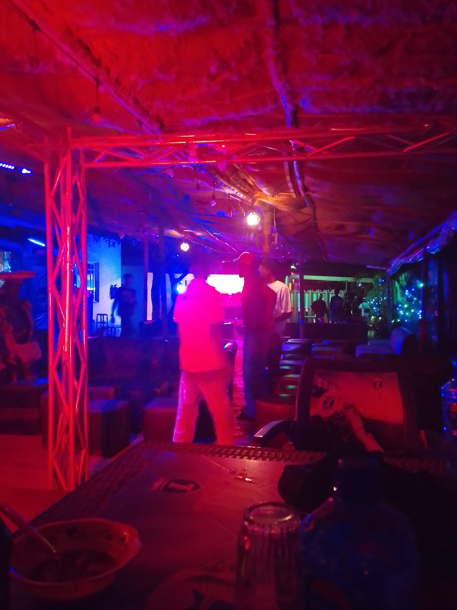 The image depicts the interior of a bar or club in an unspecified location. The lighting is predominantly red and blue, creating a dark and vibrant atmosphere. Several people are present, engaged in conversation or standing around. The foreground features a table with a bowl containing food and a glass. The background shows more seating arrangements and other individuals, suggesting a social gathering is taking place. The ceiling is partially obscured by a structure with lighting fixtures, adding to the overall ambiance of the venue.