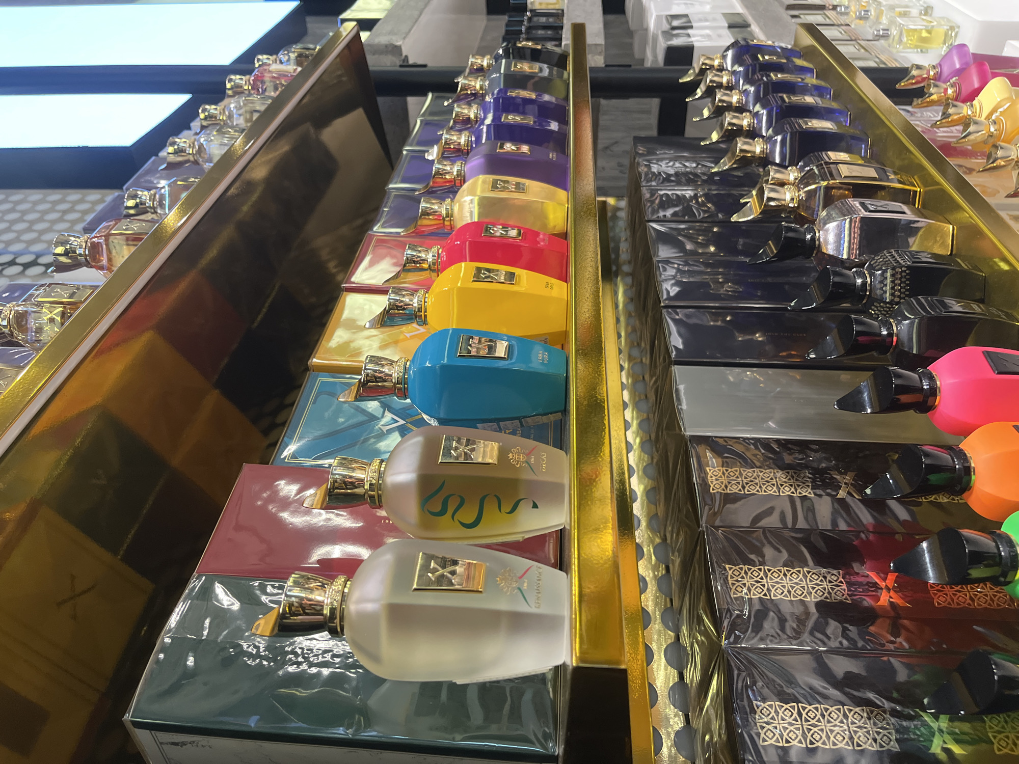 The image displays a shelf in what appears to be a retail setting, most likely a fragrance or perfume shop. Rows of perfume bottles of varying colors and designs are arranged on shelves. The shelf design consists of a gold-toned metal frame. The perfumes are arranged in a gradient of color, starting from lighter shades and progressively transitioning into deeper hues.
Based on the "X" logo on some of the boxes and the "Renaissance" label on one of the bottles, it is highly likely the perfumes are from the brand Xerjoff.
It is impossible to determine the specific location within the city of Sofia, Bulgaria, from the provided image alone.