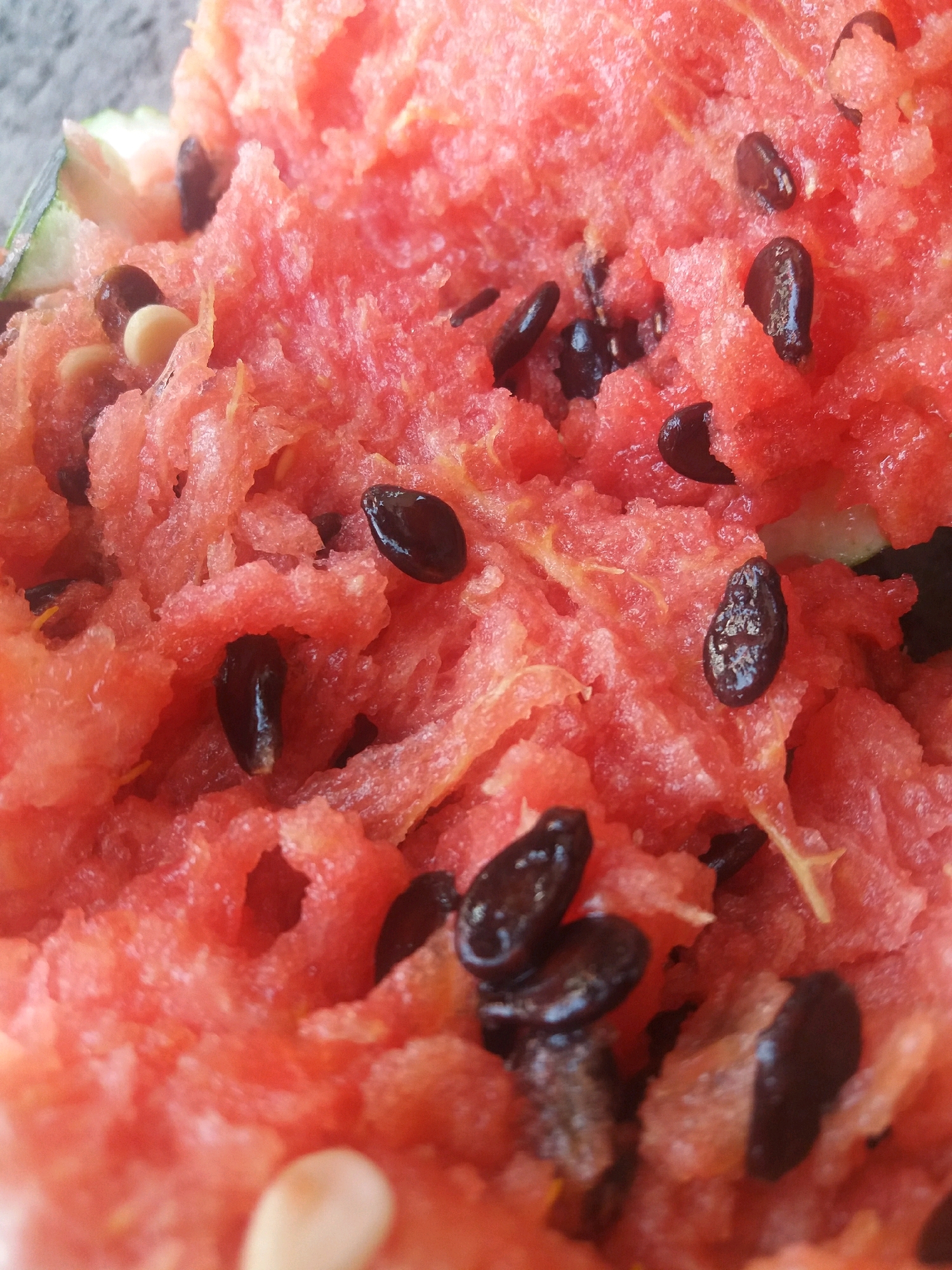 This is a close-up, top-down, eye-level shot of a fragmented watermelon. The watermelon flesh is a vibrant pink, with a shredded texture, and contains numerous black seeds scattered throughout. The background is blurred and appears to be a light gray, possibly a surface. There are no people or discernable environmental features of Dovzhyk, Ukraine present in the image. The image focuses solely on the texture and details of the watermelon.