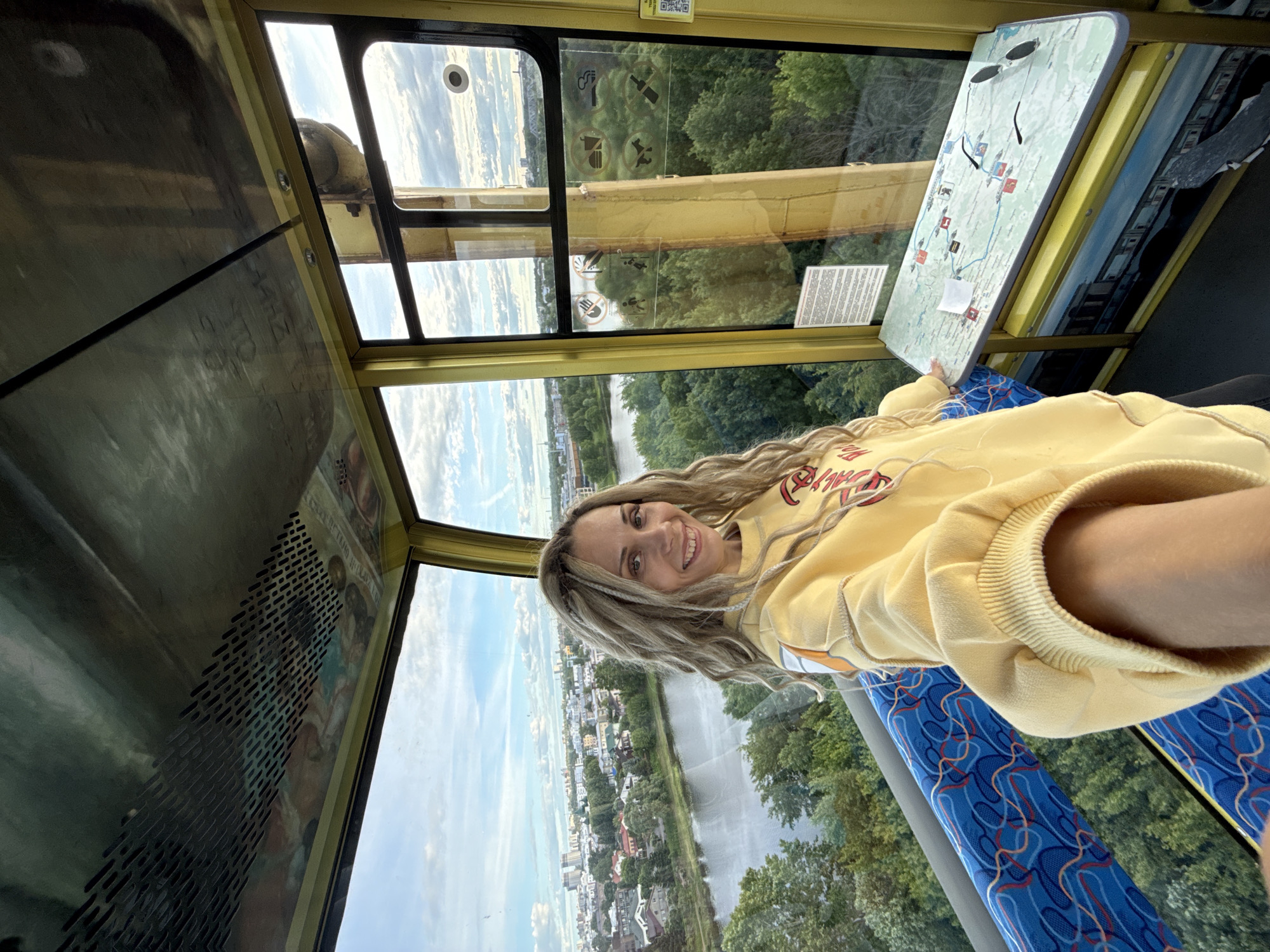 The photograph shows a woman with long, blond hair and a yellow sweater seated inside a cable car. The cable car has large windows offering views of the city below, including buildings, a river, and dense foliage. Inside the cable car, there's a map or informational display attached to the side, and blue patterned seating. The photo is taken from an unusual angle. Based on the general architecture and layout, the location might be in or near the city of Шебунино, Russia.