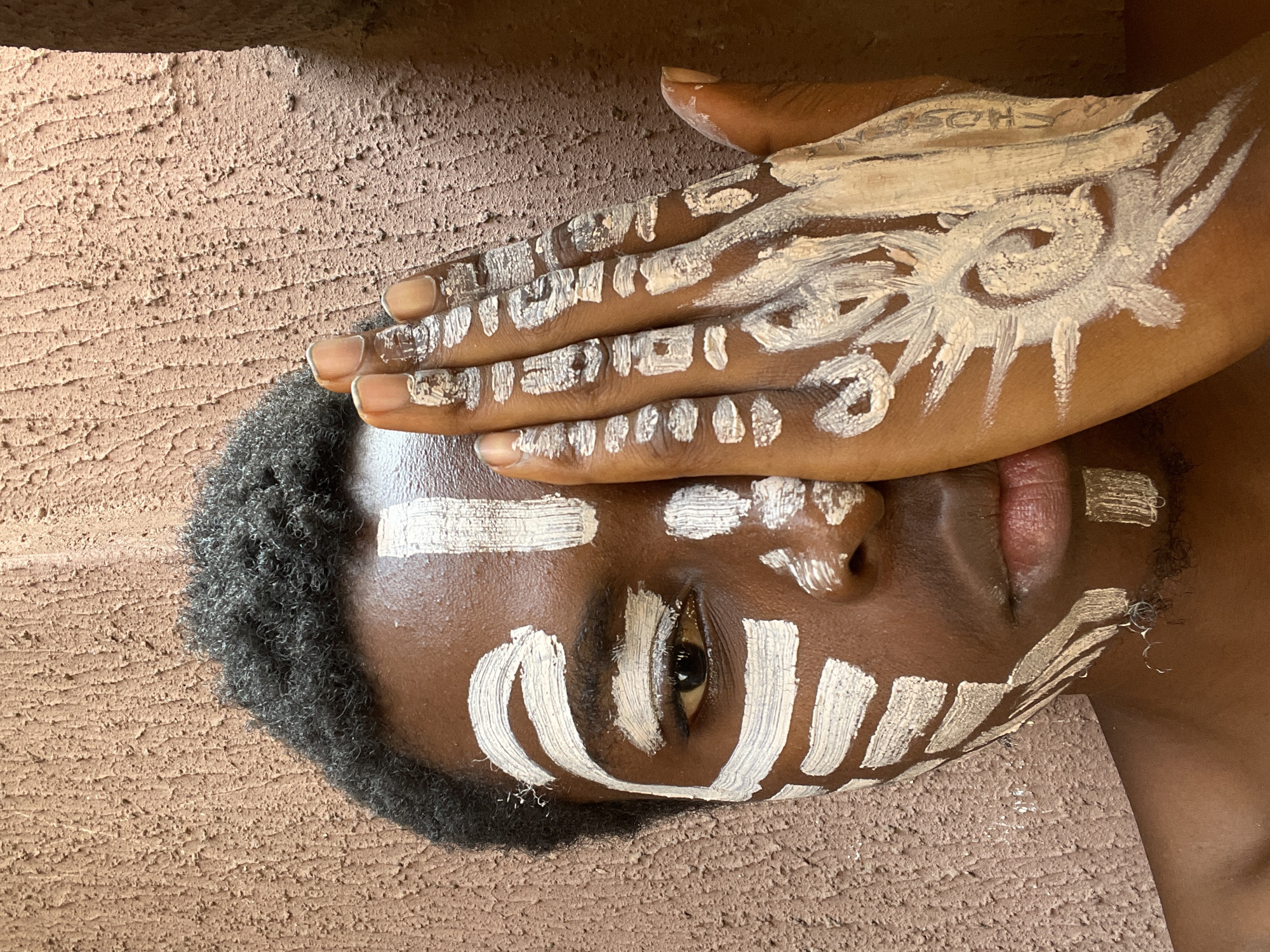 A close-up, sideways-oriented portrait features a person with dark brown skin, adorned with white body art on their face and hand. The subject's head rests against a textured, reddish-brown surface, likely a wall, suggesting a setting in Onitsha, Nigeria.
The face is intricately decorated with bold, white stripes: a horizontal band across the forehead, shorter vertical and curved marks around the visible right eye, and U-shaped patterns on the cheek and chin. The eye itself is dark brown and directly engages the viewer. Dark, short, curly hair frames the side of the head, blending into the background.
The person's right hand is placed over the upper part of their face, partially obscuring the left eye and forehead. This hand is also painted with white patterns, including a prominent sun-like or starburst symbol with a spiral center on the back of the palm. The fingers and knuckles bear smaller, dotted or abstract white markings. Faint, possibly pre-existing text reading "CHOSEN" is discernible beneath the paint on the upper part of the hand. The nails are natural and unpainted. The lips are visible, appearing naturally pinkish.
The overall scene conveys a sense of cultural expression, artistic adornment, or ceremonial preparation. The coarse, earthy texture of the background provides a rustic context for the detailed body art.