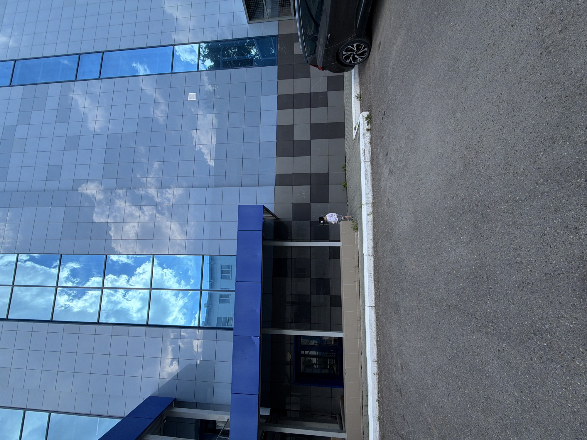 The image captures the facade of a modern building with a blue-tinted glass exterior reflecting a cloudy sky. The building features a horizontal band of blue material above the ground floor, which is a darker, checkered pattern of gray and black panels. To the right of the building, a dark-colored minivan is parked. In front of the building, on the sidewalk, a young child wearing a light-colored outfit stands with their back to the camera, seemingly looking at the building or the sky. The foreground is an asphalt surface, suggesting a street or parking lot, with a white concrete curb separating it from the sidewalk. The overall scene is urban, with a clear emphasis on the architectural design of the building and the presence of a single pedestrian. The location is specified as Chaikovsky, Russia.