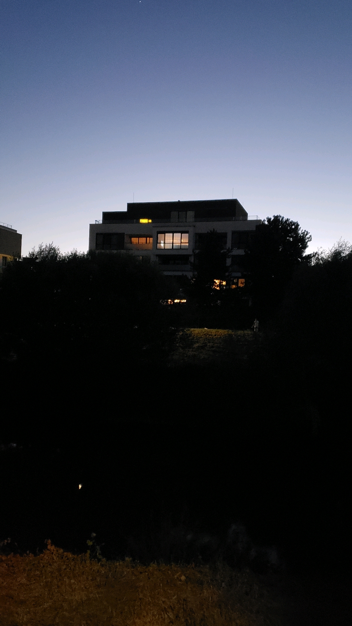 This is a low-light, vertical, medium shot of a modern building at dusk or night, with trees and vegetation in the foreground. The sky is a gradient of dark blue to lighter blue near the horizon. The building, which appears to be an apartment complex or office building, is silhouetted against the sky, with several windows illuminated with warm yellow light. Trees and dark foliage obscure the lower portion of the building and the immediate surroundings. In the foreground, there is a dark, undefined area with some texture suggesting dry grass or brush. A faint, solitary light source is visible in the darkness in the lower left quadrant of the image. The image does not contain any identifiable landmarks or context that would suggest a specific location within the city of Prešov, Slovakia.