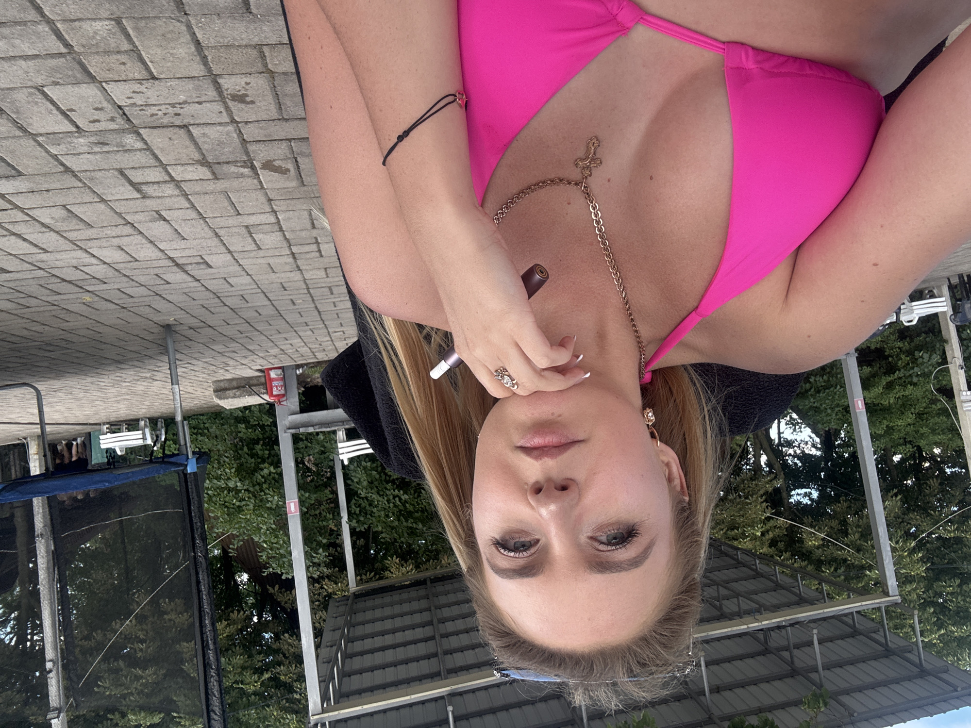 A young woman with blonde hair is captured in a selfie while sitting outside on a paved surface. She is wearing a bright pink bikini top and a gold chain necklace with a cross pendant. Her left hand rests on her chin, and she is holding what appears to be a vape pen. The background shows trees, a covered structure, and a trampoline with a blue mat and black netting. The setting suggests an outdoor recreational area, possibly near water, given the covered structure which resembles a boathouse or dock shelter.