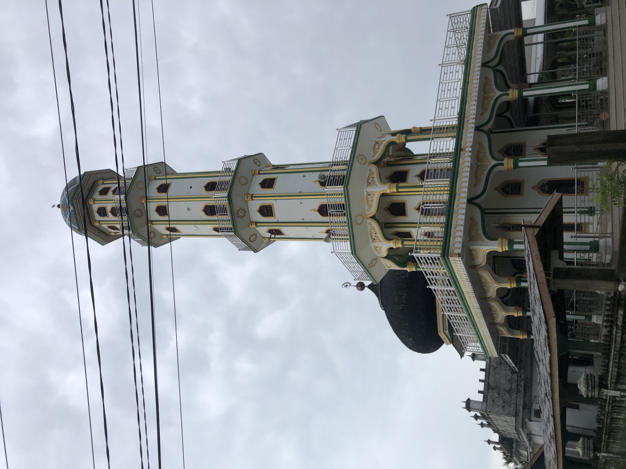The image displays a tall, ornate minaret, which is part of a mosque, set against an overcast sky. The minaret is primarily white with distinctive green and gold decorative patterns, featuring multiple levels with small windows and balconies enclosed by metal railings. A small, blue-tiled dome or cupola tops the minaret. To the right of the minaret, a large, dark-colored mosque dome is visible. Several dark power lines crisscross the upper left and central portions of the frame, partially obscuring the minaret.
The sky is cloudy and grey, indicating an overcast day. No people or specific activities are depicted; the scene is a static architectural view. The overall aesthetic of the mosque, with its minaret and dome, is consistent with Islamic architecture found in Indonesia. The specific location provided is Bale, Indonesia. There is no visible text in the image.