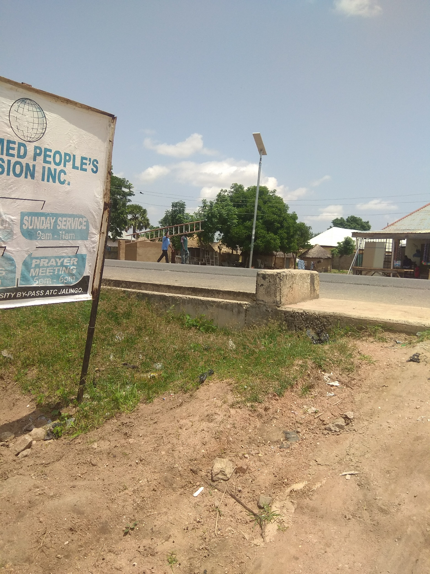 The image shows a street scene in Jalingo, Nigeria. In the foreground, there is a partially visible white banner with blue text that reads "MED PEOPLE'S SION INC." Below this, it lists "SUNDAY SERVICE 9am - 11am" and "PRAYER MEETING 5pm - 6pm". The banner also states "SITY BY-PASS ATC JALINGO".
The street is paved with asphalt and there is a concrete drainage channel along the side. In the middle ground, two men are walking on the side of the road, one carrying a ladder. There are several buildings in the background, including some with corrugated metal roofs and others with thatched roofs. A lamppost is visible on the left side of the image. The sky is clear with a few scattered clouds. The environment appears to be a semi-urban or rural area, typical of parts of Jalingo, Nigeria.