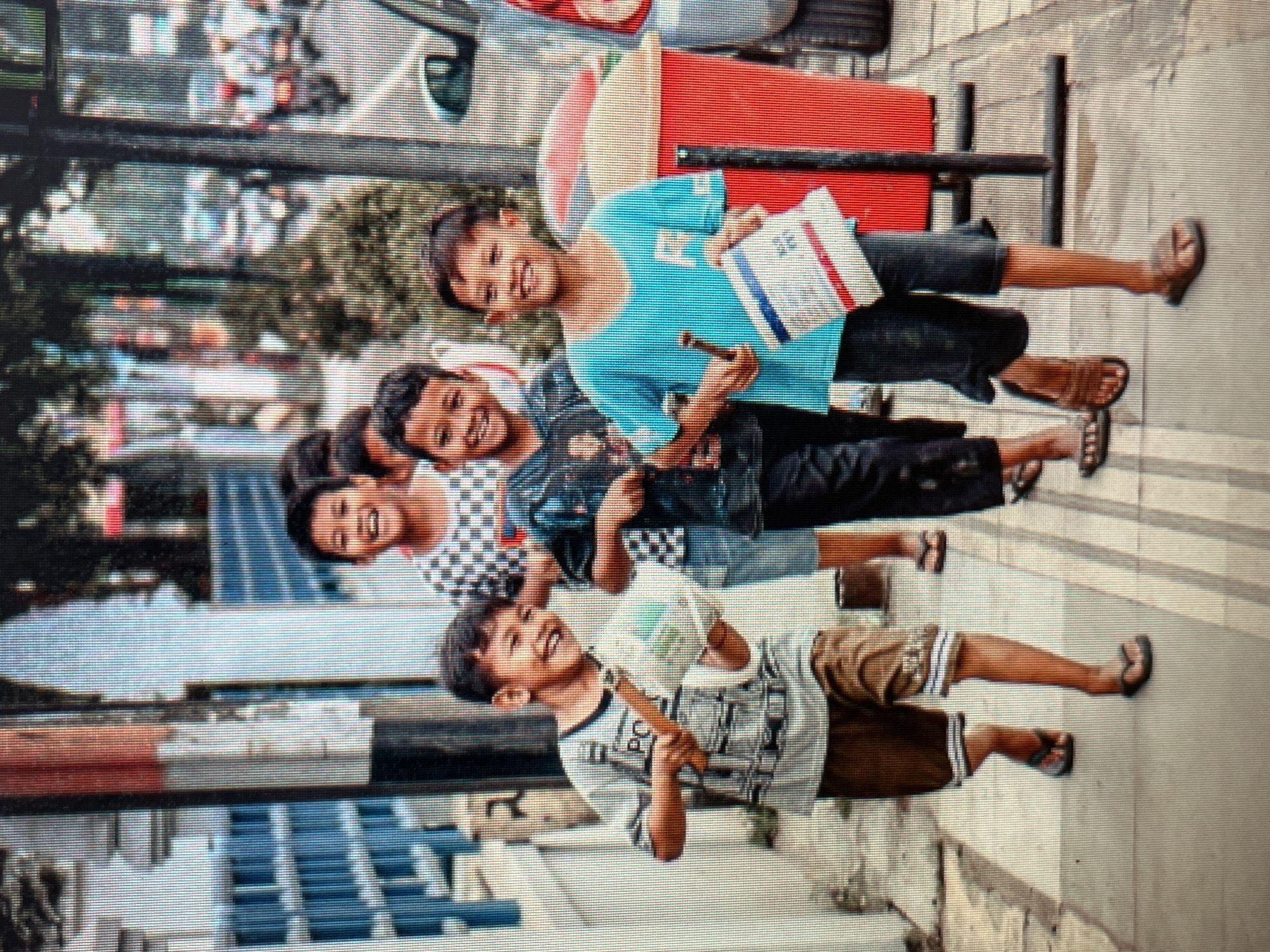 Four young boys are walking on a sidewalk in Sejahtera, Indonesia. The boys are smiling and appear to be having fun. The boy on the far left is wearing a gray t-shirt and brown shorts, and is carrying a toy gun and a bag. The boy next to him is wearing a black and white checkered t-shirt and dark shorts. The third boy from the left is wearing a black t-shirt and dark pants. The boy on the far right is wearing a blue t-shirt and dark shorts, and is holding a flag. In the background, there are trees, buildings, and motorcycles. A red trash can is also visible on the sidewalk. The overall scene suggests a lively and cheerful atmosphere in a city setting.