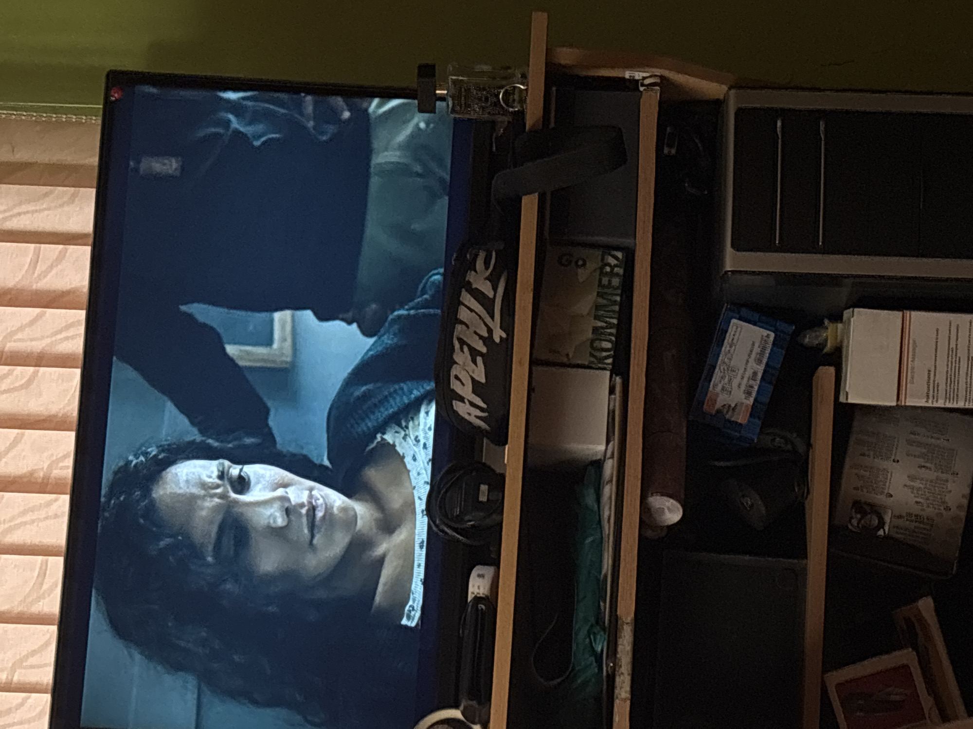 A television screen displays a scene from a film or show, depicting a woman with dark, curly hair looking downward with a distressed expression. A person's arm, clad in a dark garment, is visible behind her, appearing to hold her head or shoulder. On a shelf below the television, several items are present: a black toiletry bag with white lettering that reads "APEITZ", a remote control, a dark cord, a bottle of perfume, and a white box with text. A second shelf below holds various objects including a green plastic bag, a white box, and a black computer tower. The scene appears to be set indoors, with beige vertical blinds visible in the background of the television screen. There is no indication of the city of Brenitsa, Bulgaria in the image.