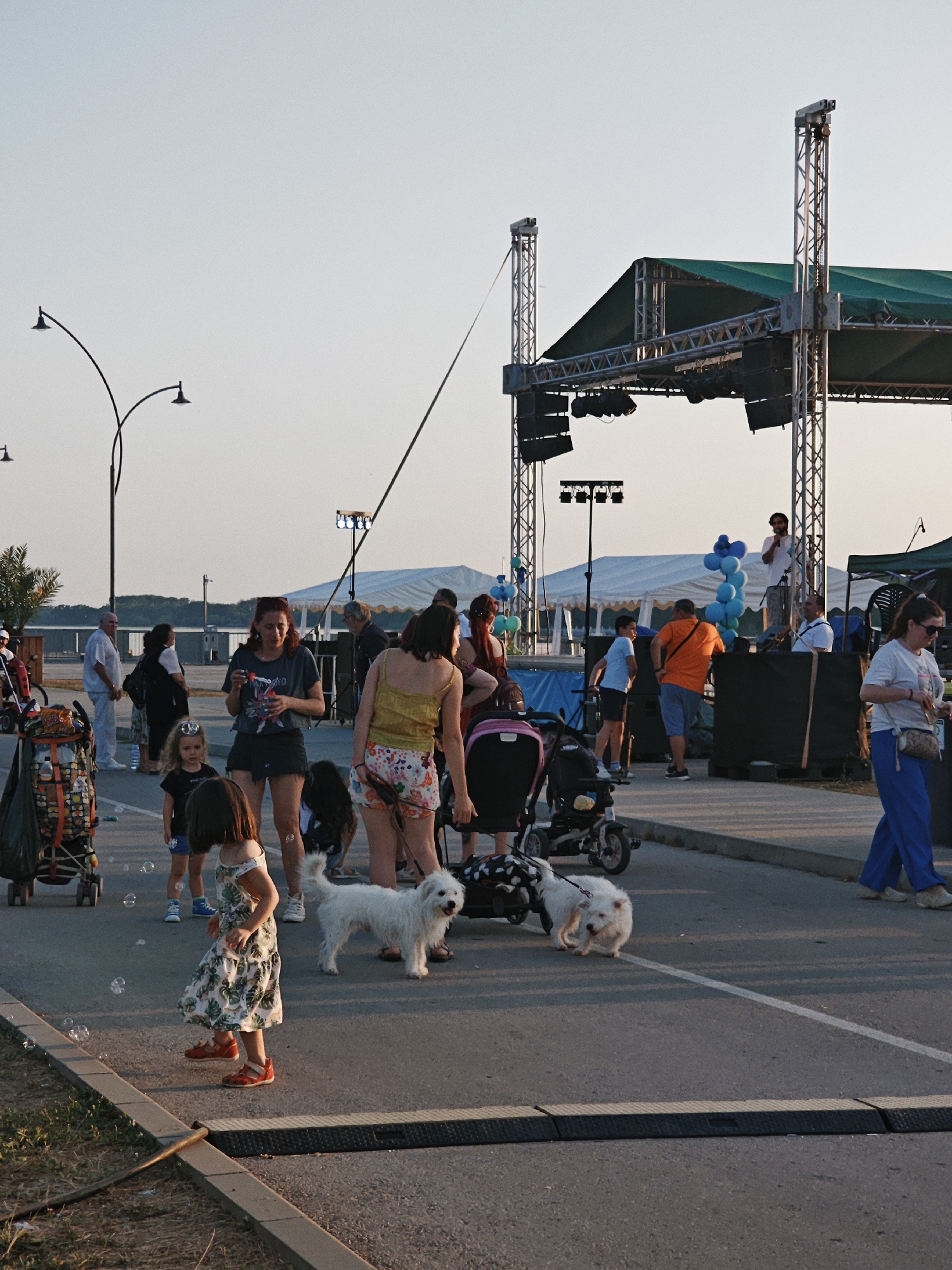 This image depicts an outdoor event with a stage set up in the background, likely for a performance or gathering. Several people of various ages are present, some walking, others with children. Two small white dogs are also visible, one on a leash. The ground is paved, suggesting an urban or park setting. The lighting indicates it is late afternoon or early evening, with a soft, warm glow. The sky is clear, hinting at pleasant weather. Bubble wands and floating bubbles suggest a playful atmosphere, possibly a family-friendly event. The overall mood appears relaxed and casual.
