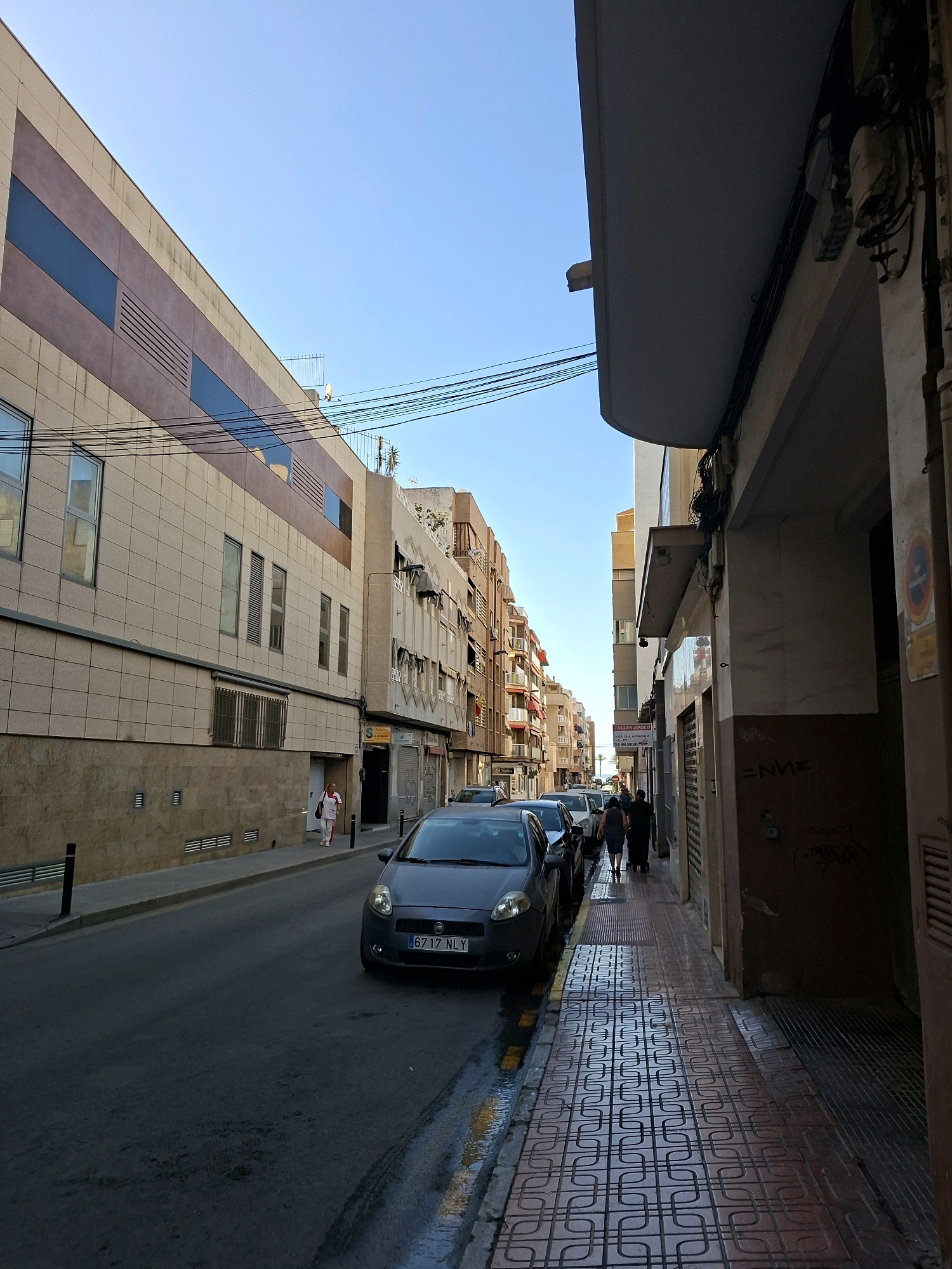 The image shows a street scene in Torrevieja, Spain, likely in the afternoon given the lighting. On the left is a multi-story building with a cream-colored facade, featuring bands of brown and blue elements, and rows of windows. A woman in white trousers and a white top with a red trim is walking on the sidewalk. To the right of the street, several cars are parked. The foreground features a dark gray car with a license plate reading "6717 NLY." The sidewalk is paved with patterned tiles, and on the right, another building features a brown facade with graffiti. Further down the street, more buildings with balconies and awnings are visible, as are other cars and pedestrians. The sky is clear and blue. Electrical wires are strung overhead.