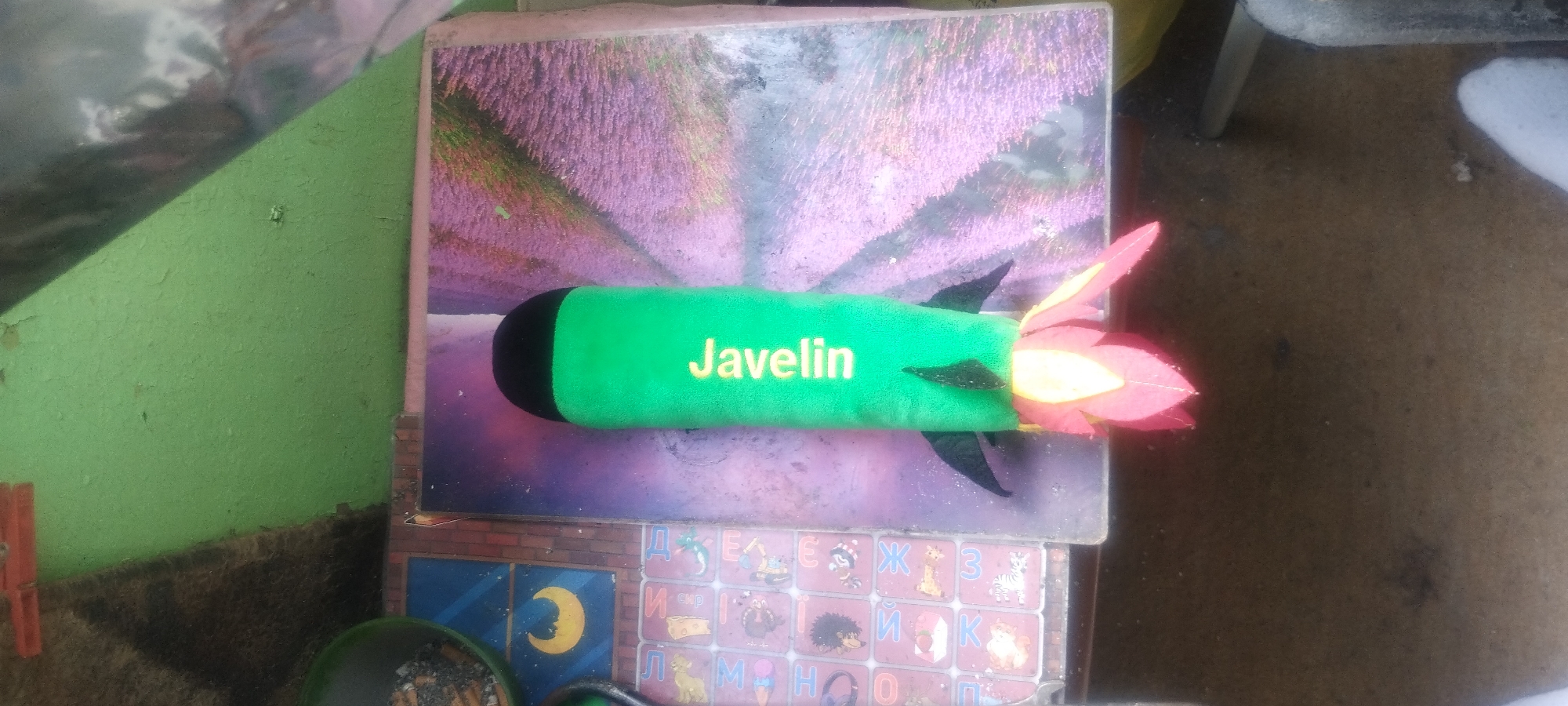 A green, plush toy rocket with the word "Javelin" printed in yellow text is positioned on top of a book featuring a photograph of a lavender field. The rocket has a black tip and what appear to be stylized flame-like fins made of red, yellow, and possibly pink material at its rear. The book is placed on a surface that includes what looks like children's educational tiles depicting letters, numbers, and animals. The background shows a green, textured wall and a brown, possibly dirt or concrete surface. The scene appears to be indoors or in a sheltered outdoor area, given the presence of the book and toy, and the lighting suggests daytime. There are no people visible. The overall impression is of a handmade or toy item displayed in a somewhat informal or cluttered setting.