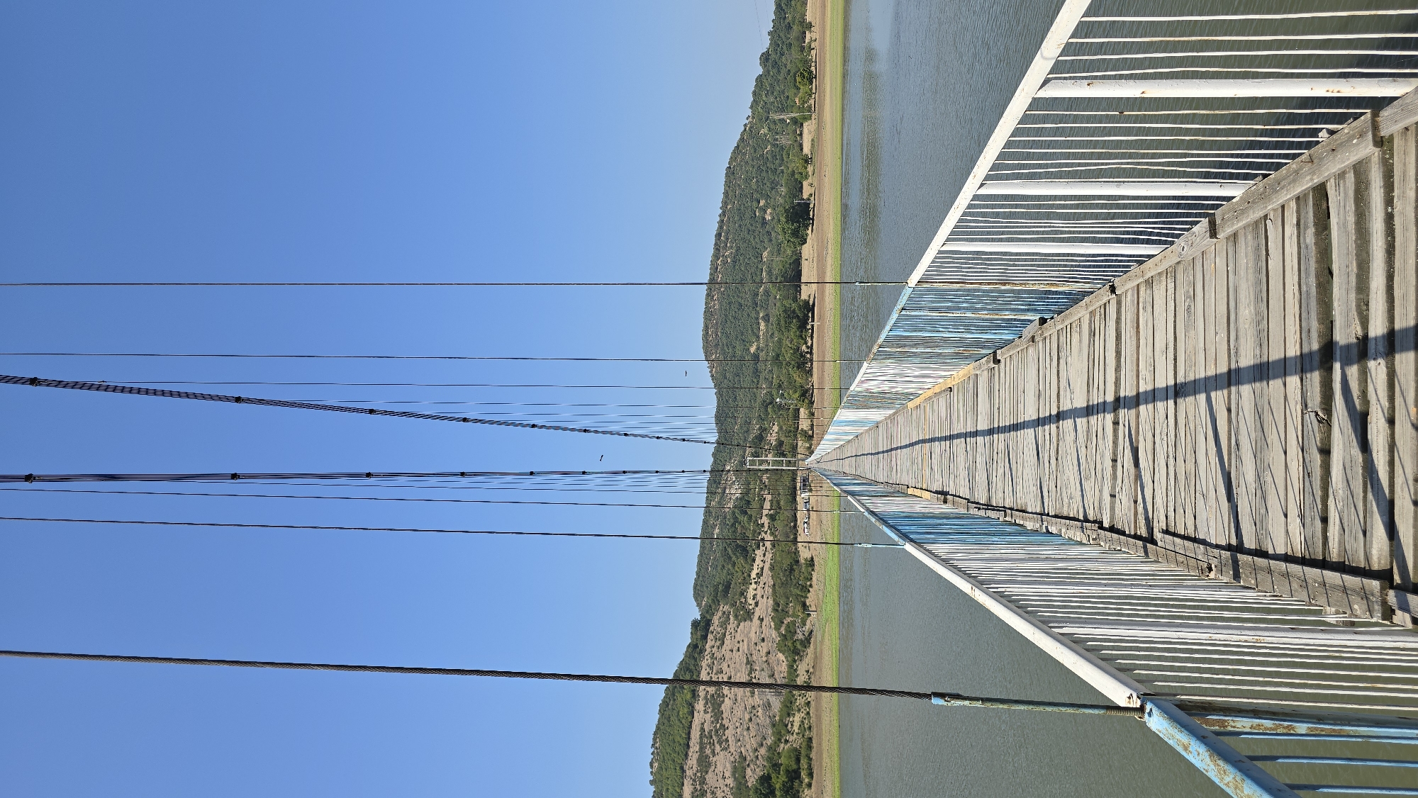 A long, narrow pedestrian suspension bridge made of weathered wooden planks and metal railings spans across a body of calm, green water. The bridge has white railings with intermittent blue accents. The bridge leads towards distant, rolling hills covered in dry vegetation and scattered trees under a clear, bright blue sky. The sun casts long shadows across the wooden walkway, indicating it is likely midday. The scene suggests a rural or semi-rural environment, possibly near the village of Lisitsite, Bulgaria, given its hilly terrain and body of water. There are no people visible on the bridge or in the immediate vicinity.
