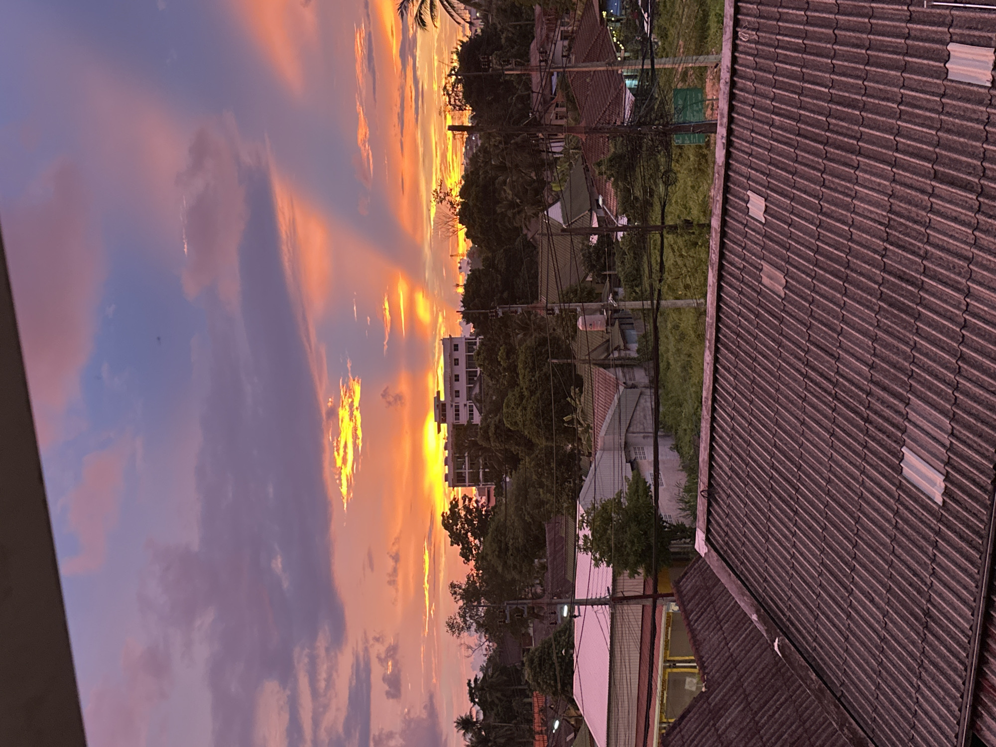 This image captures a sunset over Karon, Thailand, with a dramatic sky filled with vibrant orange, yellow, and purple hues. The sun's rays are visible as they pierce through the clouds, creating a striking visual effect. In the foreground, the tiled roofs of houses are visible, some with metal sheeting. Beyond the rooftops, lush green trees and vegetation create a dense canopy. A multi-story building with a white facade stands out amongst the trees, suggesting a built-up area within the town. Utility poles and wires crisscross the scene, indicating the presence of infrastructure. The overall impression is one of a serene, tropical evening in Karon, with the natural beauty of the sunset contrasting with the urban landscape.