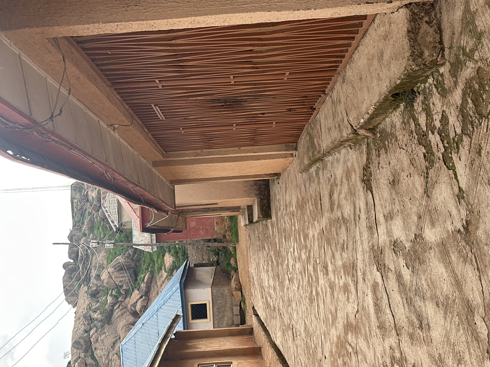 The image shows a narrow, cracked, concrete pathway in front of several buildings. On the right, a tan-colored building with a red-trimmed roof features a tall, vertical, orange metal gate. To the left, a light brown building with a dark window and a blue corrugated metal roof is visible. Further down the pathway, a red building with a similar roof style can be seen. The background is dominated by a large, rocky, arid hillside with sparse green vegetation. Electrical wires are strung across the scene, supported by poles. The sky is overcast.