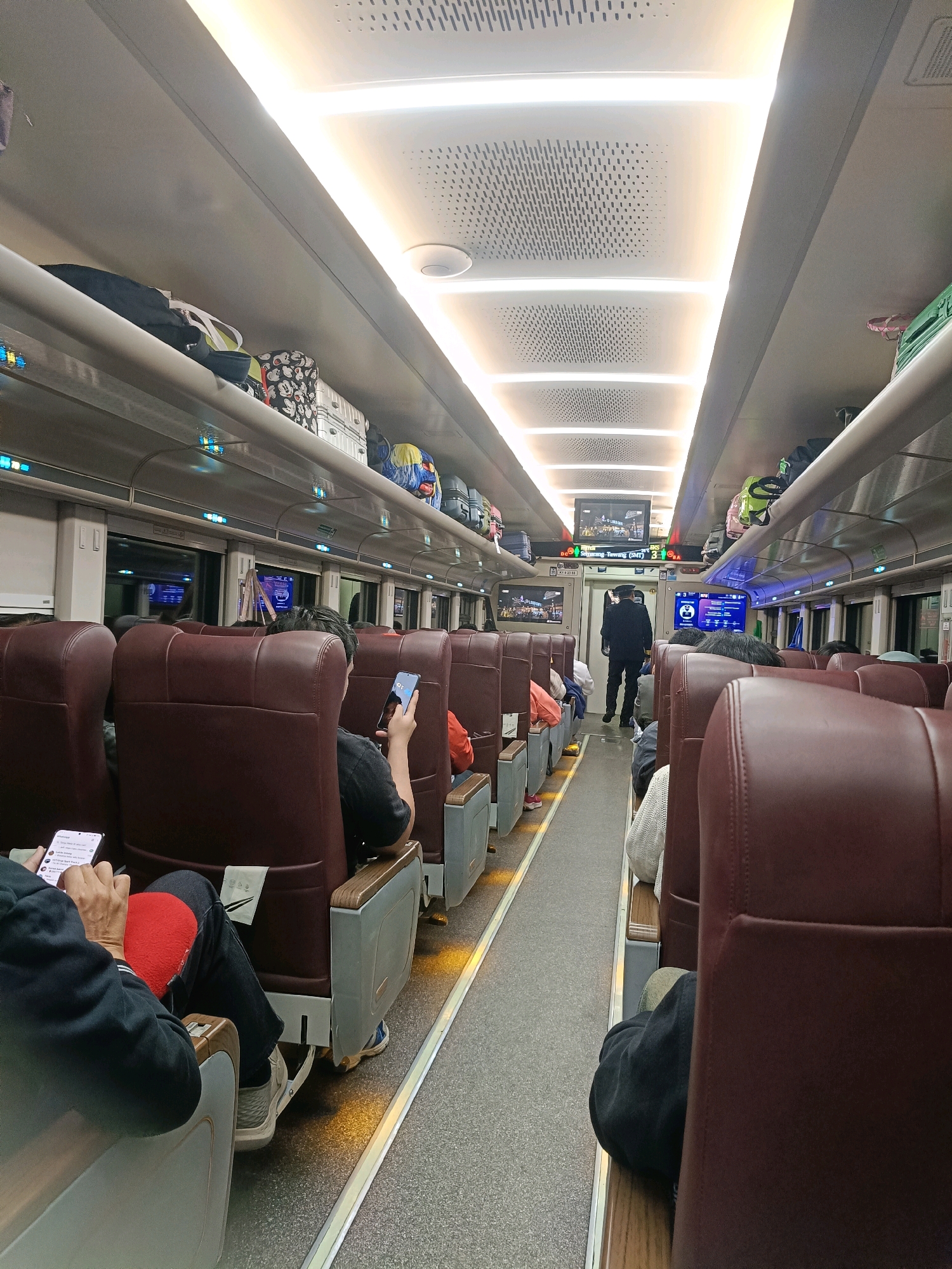 The image depicts the interior of a train carriage in Surabaya, Indonesia. The carriage features rows of plush, maroon-colored seats with wooden accents, arranged along a central aisle. Overhead luggage racks are filled with various bags and suitcases, some of which have distinctive patterns. The ceiling is equipped with modern, strip lighting that illuminates the space. In the background, a uniformed train conductor stands near a screen displaying information. Several passengers are seated, some of whom are engaged with their mobile phones. The flooring is a textured grey carpet. The overall impression is of a well-maintained and modern public transport environment.
