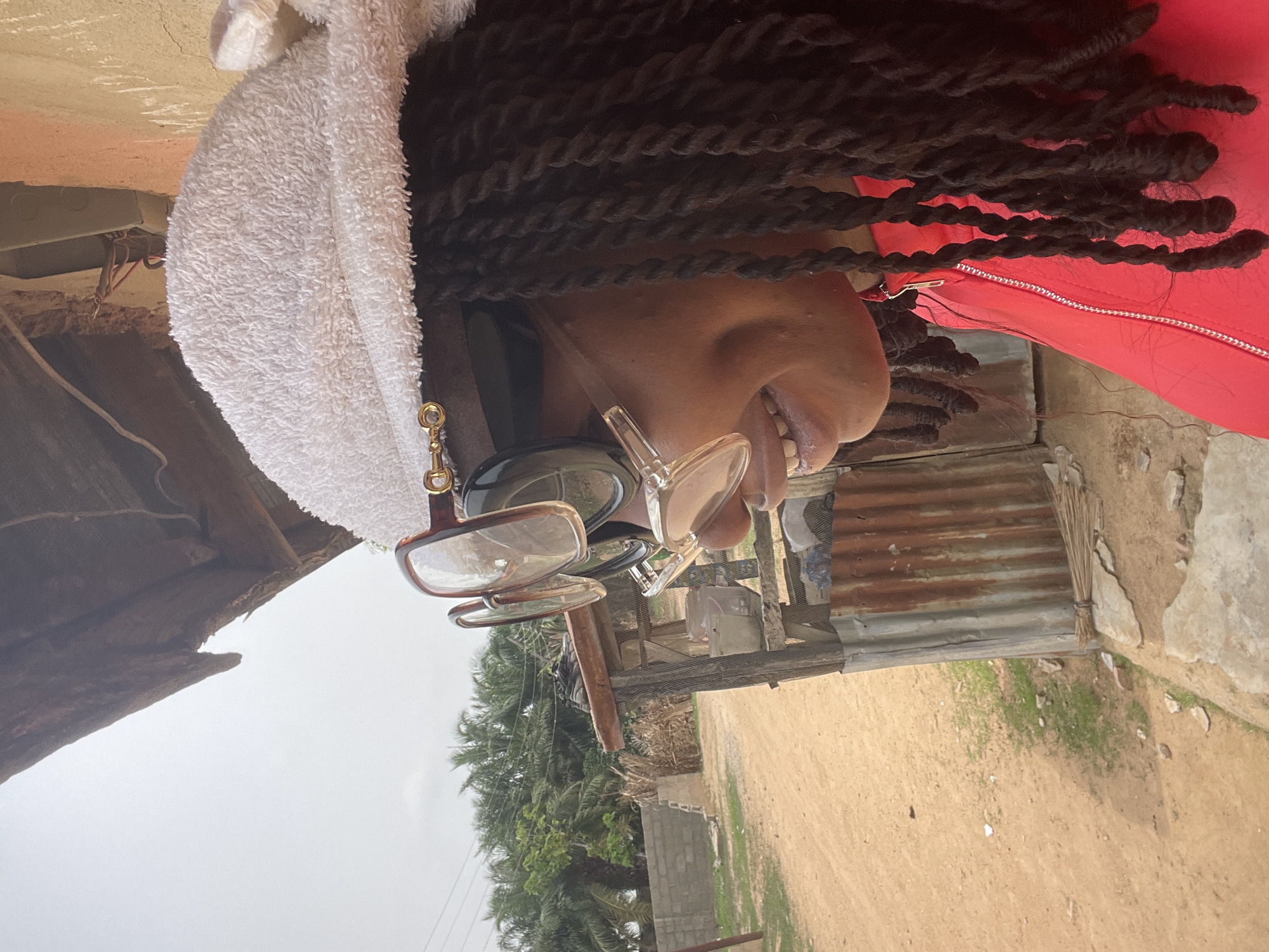 A medium shot depicts a person with dark braided hair, wearing multiple pairs of glasses and a white towel over their head, with a red garment visible. The background suggests a rural or less developed area, typical of a smaller city such as Akwanga, Nigeria. There are structures made of corrugated metal and wood, indicative of local housing or sheds. A sandy ground and vegetation, including palm trees, are also visible. The overcast sky suggests daytime.
