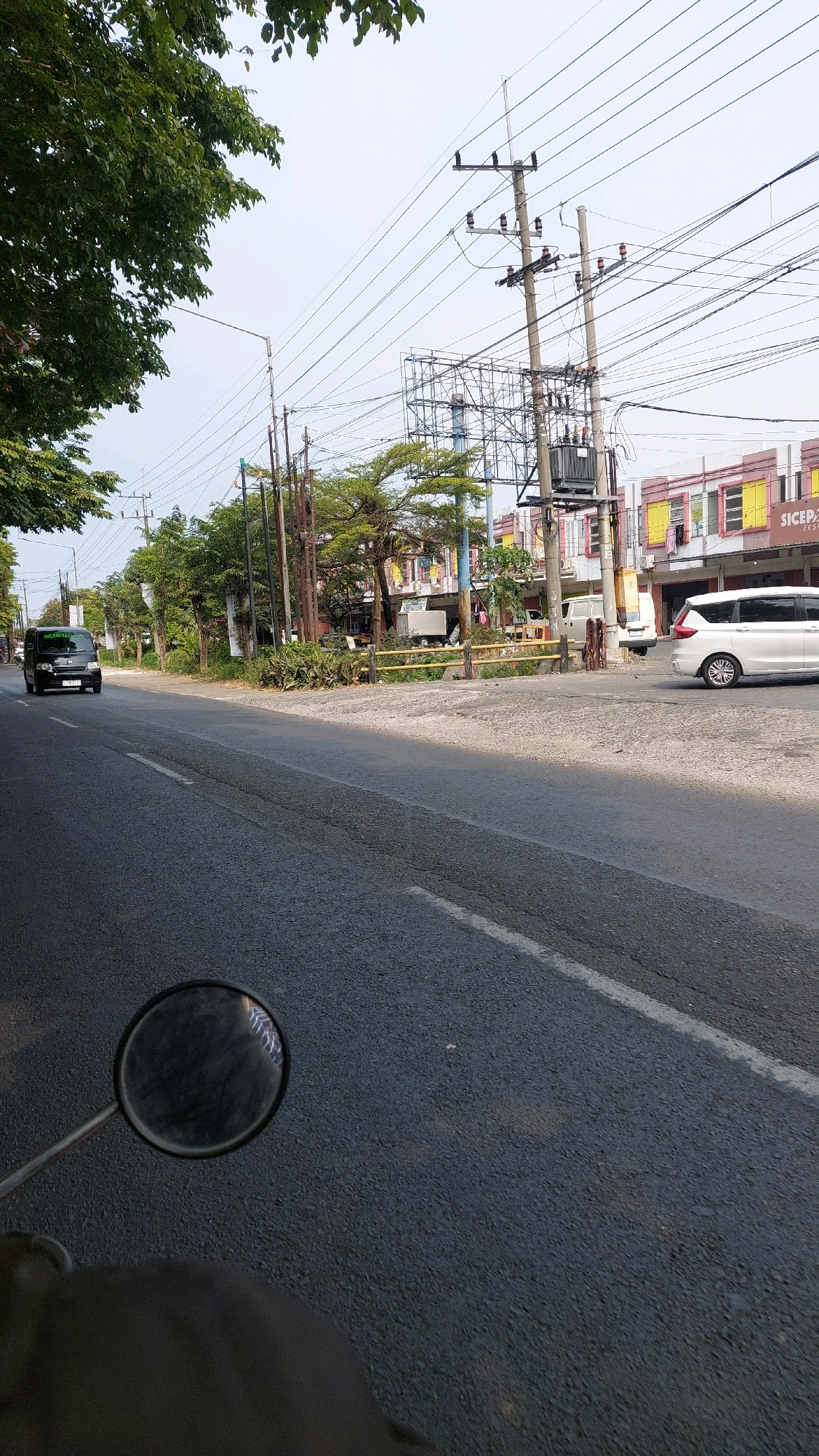 The image depicts a street scene in Ketimang, Indonesia, as suggested by the provided metadata. A black minivan with text on its windshield is traveling down the left lane of a paved road. A white SUV is visible in the right lane. The roadside is lined with trees and utility poles, with a building complex featuring yellow and pink facades in the background. A yellow barrier runs along the edge of the sidewalk. The perspective appears to be from a motorcycle, with a rearview mirror in the lower left corner reflecting the sky and surrounding structures. The sky is overcast, and the overall lighting suggests daytime.
