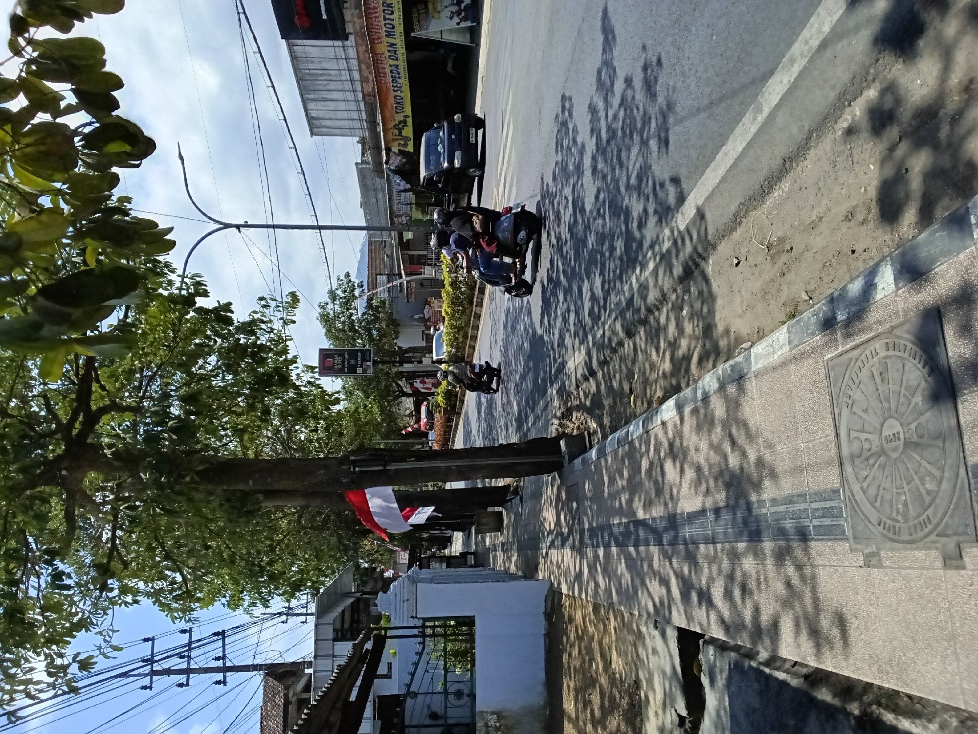 This is a photograph of a street scene in Kelutan, Indonesia, on a sunny day with a blue sky and scattered clouds. A sidewalk runs along the left side of the road, paved with square tiles and featuring a tactile paving strip in the center. A manhole cover with a circular pattern is visible on the sidewalk. Trees with green foliage are planted along the sidewalk, casting shadows on the pavement. Indonesian flags, red and white, are displayed on poles.
The road itself has two lanes of traffic. Several vehicles are present: a black pick-up truck, a scooter with two people on it, and another scooter partially visible in the foreground. The people on the scooters are wearing helmets.
Shops with signage are visible on the right side of the road, including one with the text "TOKO SEPEDA DAN MOTOR". A tall street light stands on the right side of the road, and electrical wires are strung overhead. The overall environment appears to be a commercial or residential street in a town or city.