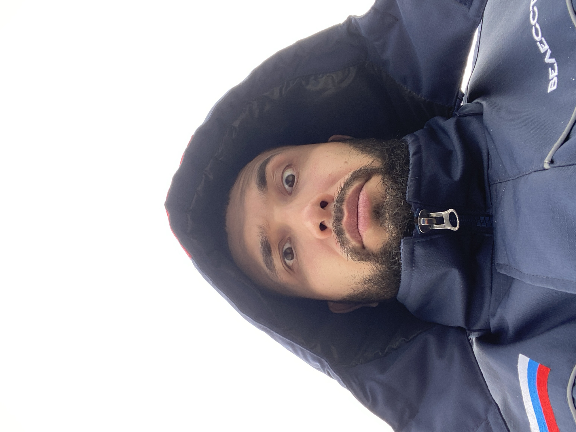 A close-up, upward-angled shot shows a man wearing a dark blue hooded jacket with a zipper. The jacket has a patch featuring red, white, and blue stripes. The man has brown eyes, a beard, and is looking slightly upward. The background is a bright white, likely indicating an outdoor scene with overcast lighting. The location is not deducible from the visual information. The man appears to be alone and no particular interaction can be determined.