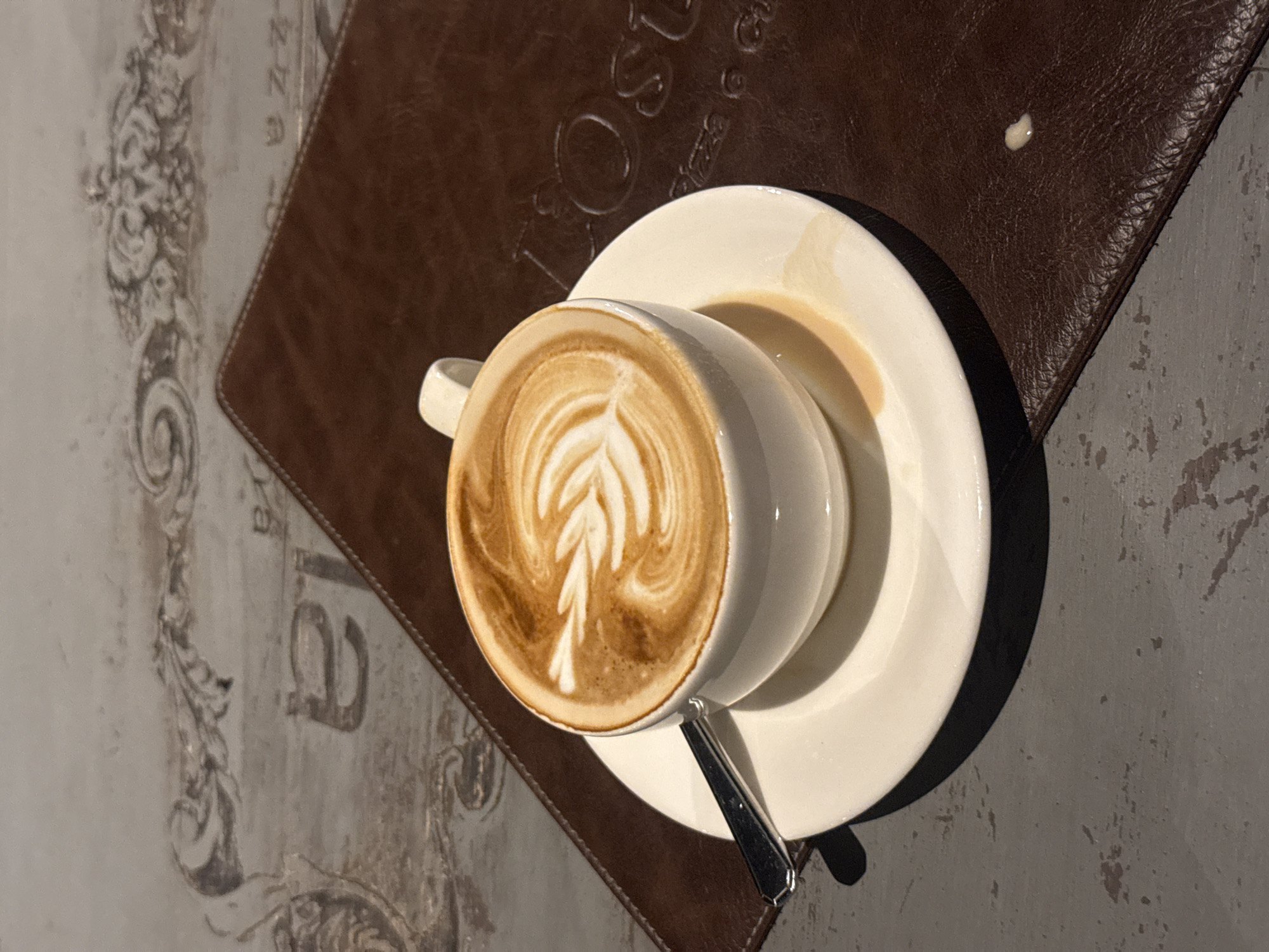 A close-up, eye-level shot captures a white ceramic cup of latte art coffee, placed on a white saucer, which in turn rests on a brown leather placemat. The placemat is positioned on a textured, grey table with visible wear. The latte art features a delicate leaf pattern formed by frothed milk. A silver spoon rests on the saucer, partially submerged by the cup. The background reveals a distressed, light-colored surface with ornamental detailing, possibly a table or wall. The overall lighting is soft, creating subtle shadows. No people are visible in the frame. The context of Denpasar, Indonesia, is not directly conveyed by the visual elements of the image.