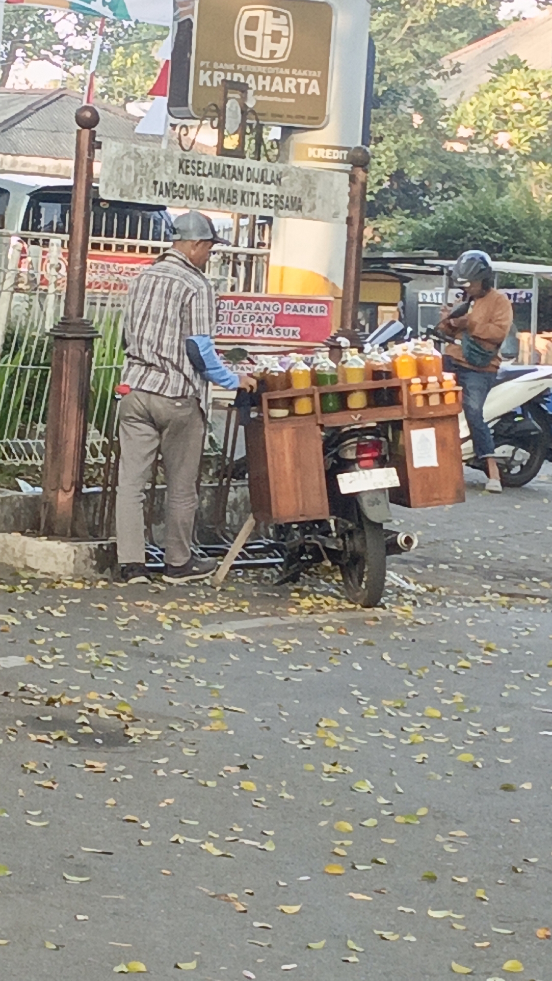The image captures a street scene in Salatiga, Indonesia, likely during the day given the lighting and shadows. In the foreground, a man wearing a striped shirt and a grey cap stands beside a motorcycle with a wooden cart attached to its rear. The cart is laden with several bottles of colorful liquids, possibly beverages. Behind him, a red sign reads "DILARANG PARKIR DI DEPAN PINTU MASUK," which translates to "NO PARKING IN FRONT OF THE ENTRANCE." Further back, another individual is seated on a motorcycle, wearing a brown jacket and a helmet. The background shows trees and buildings, consistent with an urban environment. Fallen leaves are scattered across the asphalt road, suggesting it might be the autumn season. A sign for "KRIDAHARTA" with a logo is visible on a pole, indicating a business or institution.
