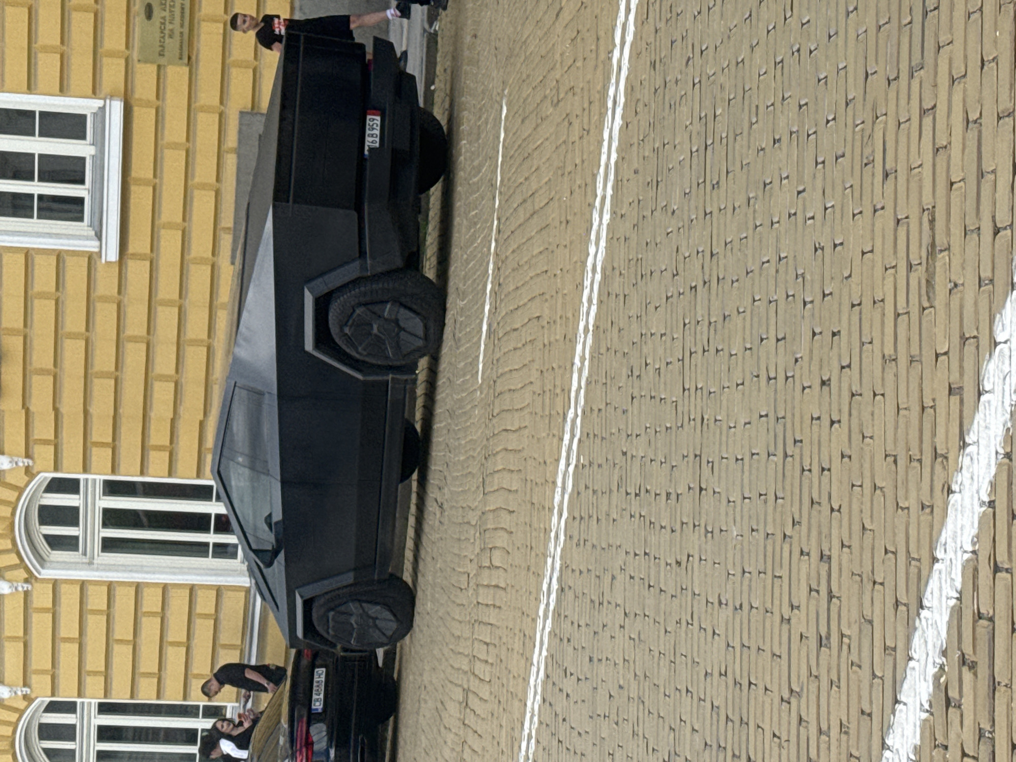 The image shows a Tesla Cybertruck parked on a brick-paved street in front of a yellow building with white windows. A person stands near the back of the vehicle. Other people are visible near the other vehicle. The Cybertruck has a license plate that reads "16 B 959" and another vehicle is near the truck with license plate "CB 4649 HO". The scene appears to be in an urban environment.