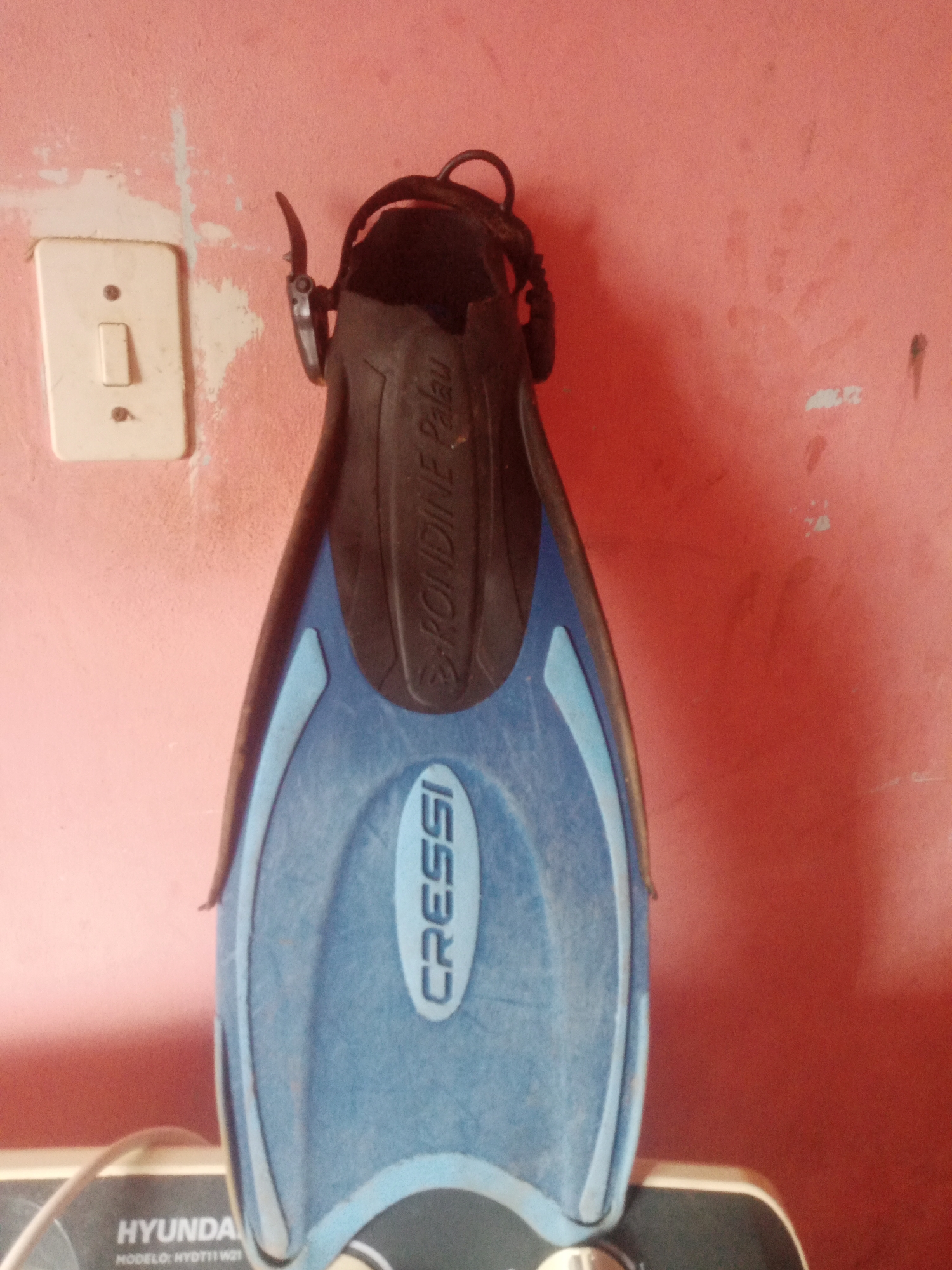 A single blue and black swimming fin with the brand name "CRESSI" is hanging vertically on a pale pink wall in La Tagua, Venezuela. The fin is oriented with the heel strap at the top. To the left of the fin, a white electrical light switch is mounted on the wall. The wall shows some signs of wear and discoloration. The bottom portion of the image reveals a black surface with the word "HYUNDA" printed in white, suggesting the fin is positioned in front of or on top of a Hyundai appliance. A white cable is visible extending from the left side of the frame.