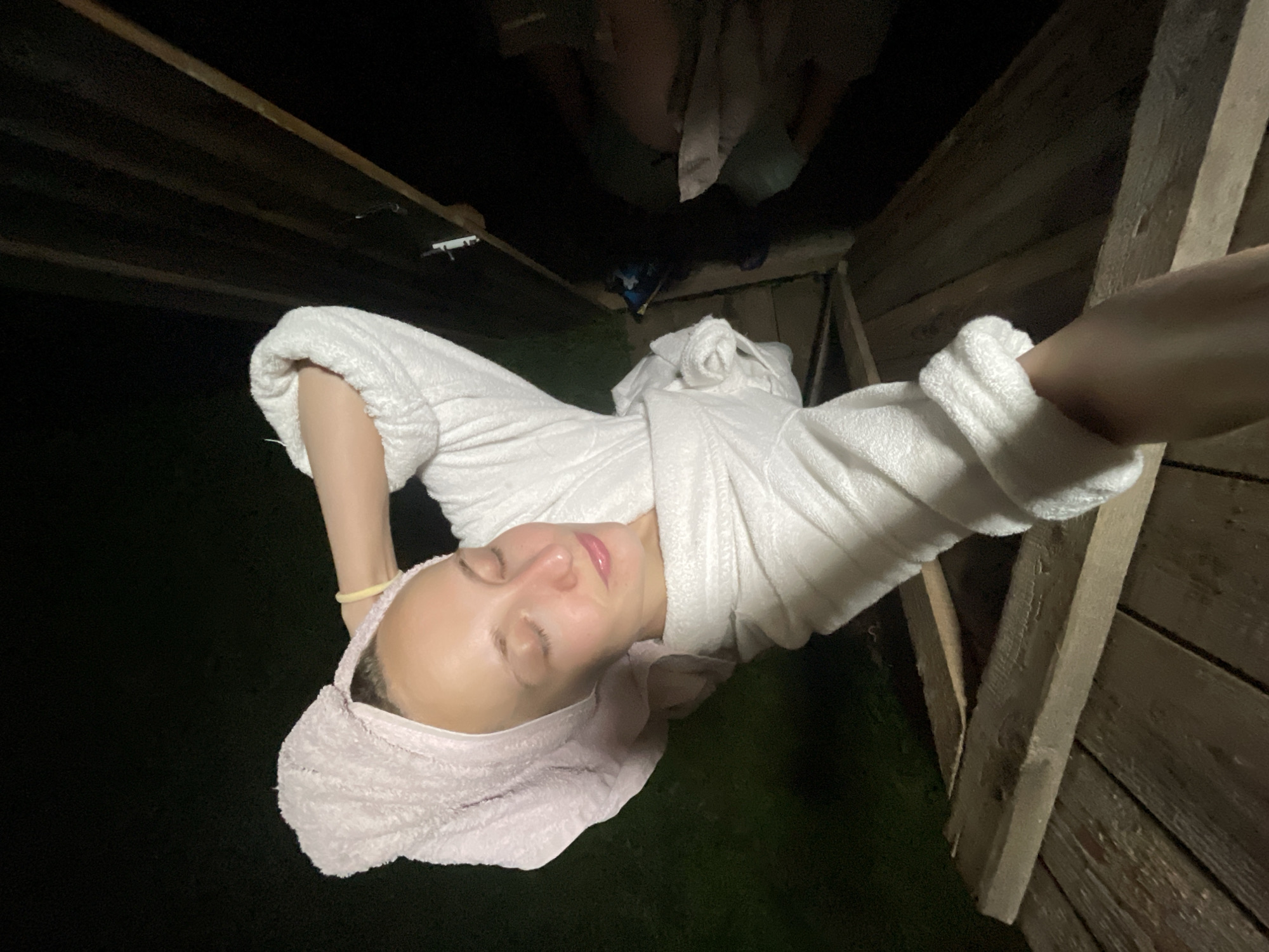 A selfie is taken from a high angle, looking down at a woman in a white bathrobe and a pink towel wrapped around her head. She is lying on a wooden structure, possibly a deck or a porch, outdoors at night. Her eyes are closed, and she has a relaxed expression on her face. Her right arm is bent and resting on her head. There is a yellow bracelet on her wrist. The background is dark, with some blurry green grass visible to the left. To the right, there are wooden planks and shadows, suggesting an outdoor setting. The overall lighting is dim, indicating it is nighttime. The location of Sharkan, Russia, is not discernible from the visual information present in the image.
