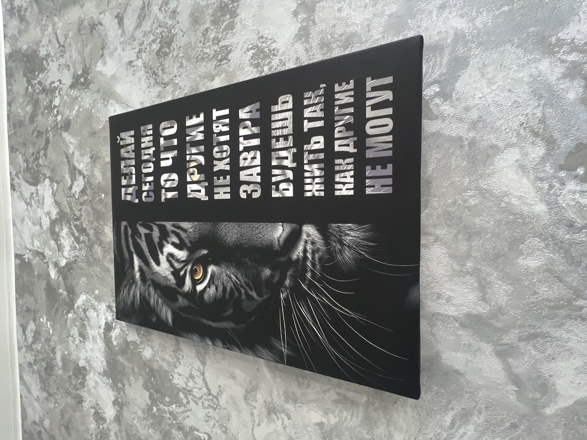 Here's a matter-of-fact description of the image:
The image shows a rectangular canvas print hanging on a gray and white textured wall. The print features a black and white image of a tiger's face, specifically the upper part of its head including one eye, and whiskers. Above the tiger image are several lines of text in a silver or metallic font. The text is in Russian and translates to "Do today what others don't want to, tomorrow you will live as others cannot". The canvas is mounted on the wall in the city of Unknown, Unknown.