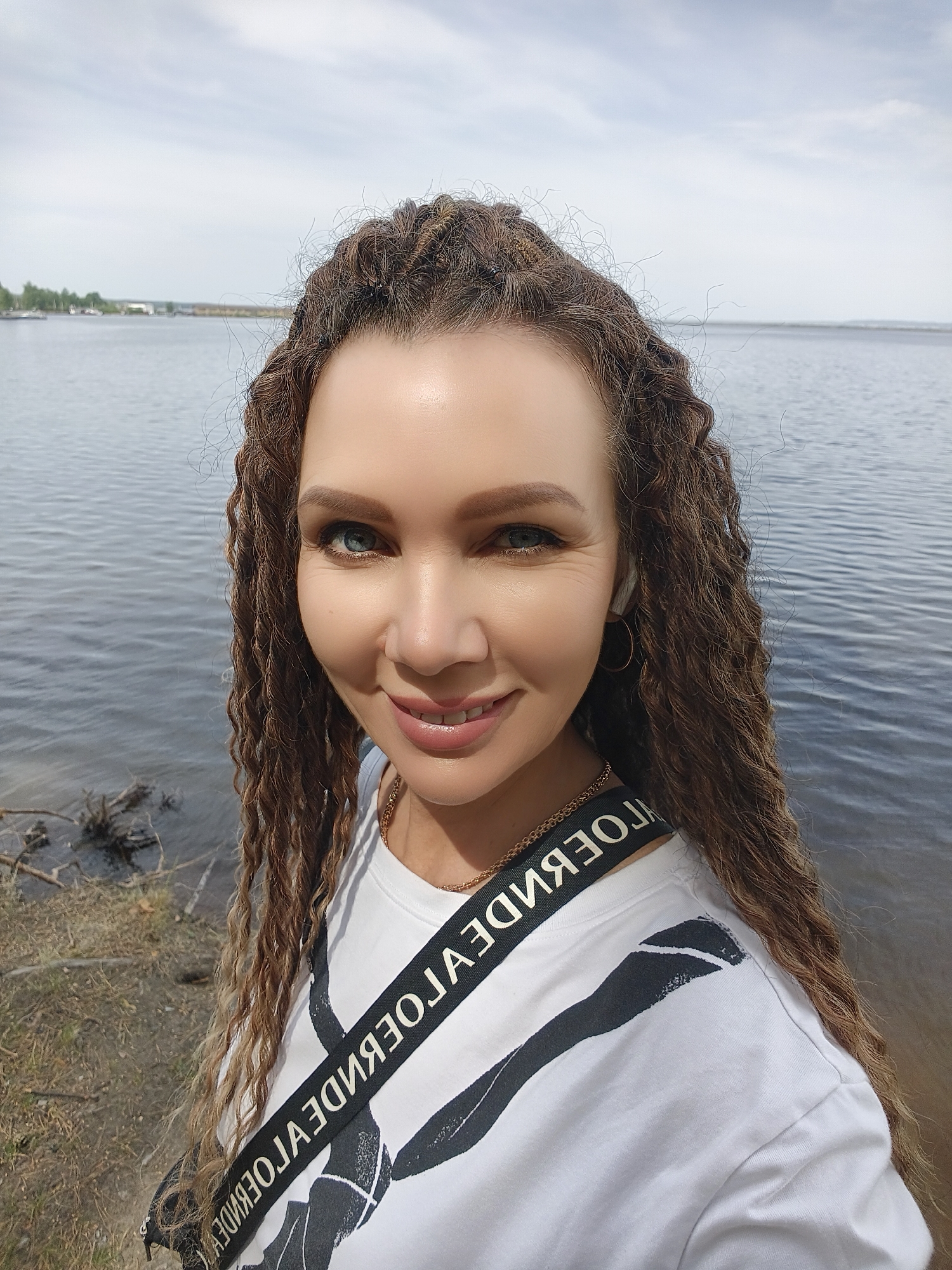 A woman stands outdoors near a body of water. The woman has long brown hair that is styled in braids and curls. She is wearing a white t-shirt with a black strap across her chest. The background consists of a lake and the sky. The location is Unknown, Unknown.