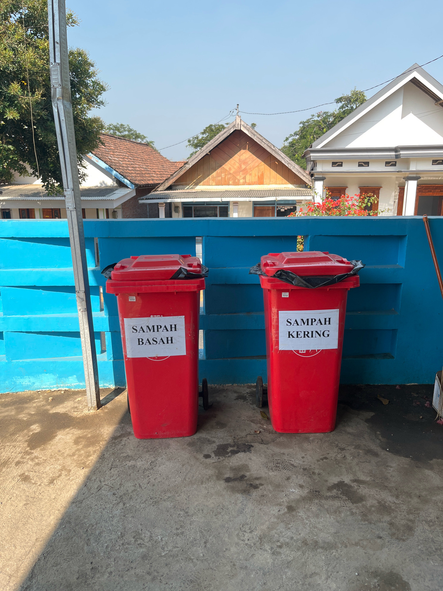 Two red trash bins with black lids are placed side-by-side in front of a bright blue fence. The bin on the left has a white label that reads "SAMPAH BASAH" (Wet Waste). The bin on the right has a similar white label that reads "SAMPAH KERING" (Dry Waste). Both bins have black plastic liners visible. The bins have wheels at the bottom. The ground in front of the bins is concrete, with shadows cast from a pole on the left. Behind the fence, there are houses with tiled roofs and some greenery, suggesting a residential or semi-residential area in Sumberjo, Indonesia. The sky is clear and blue.