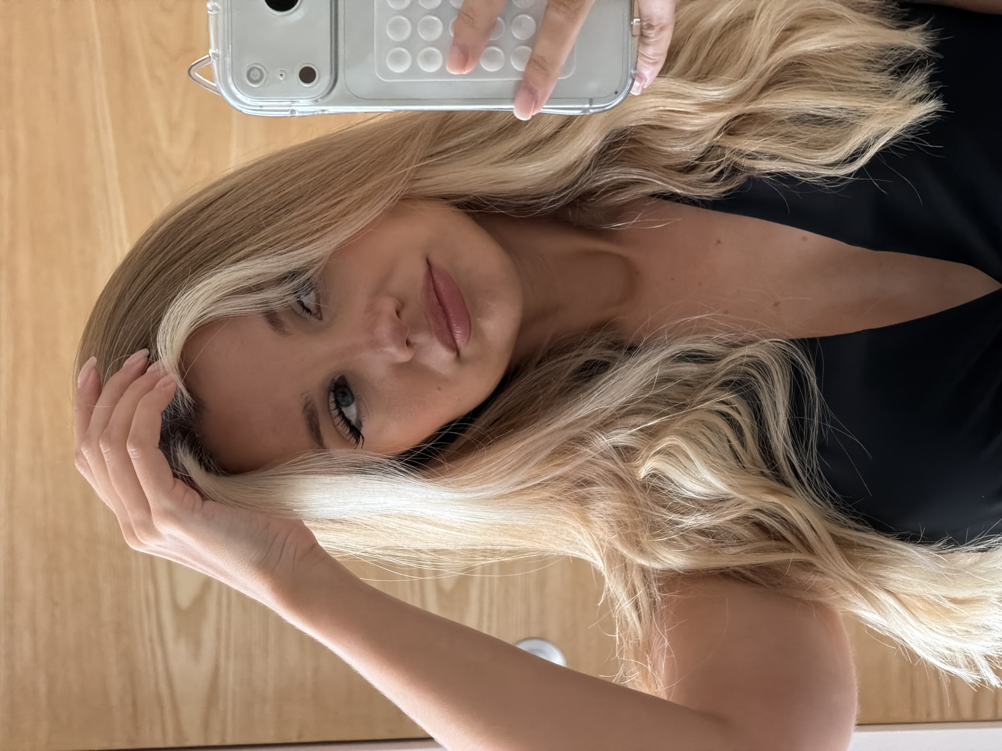 A young woman with blonde hair, featuring a prominent white streak, takes a mirror selfie. She is wearing a black, low-cut top. Her right hand is raised, touching her hair, while her left hand holds a clear phone case. The background appears to be a room with wooden paneling. The image appears to have been taken indoors, likely in a residential setting. There is no indication of the location being Thep Krasatti, Thailand.