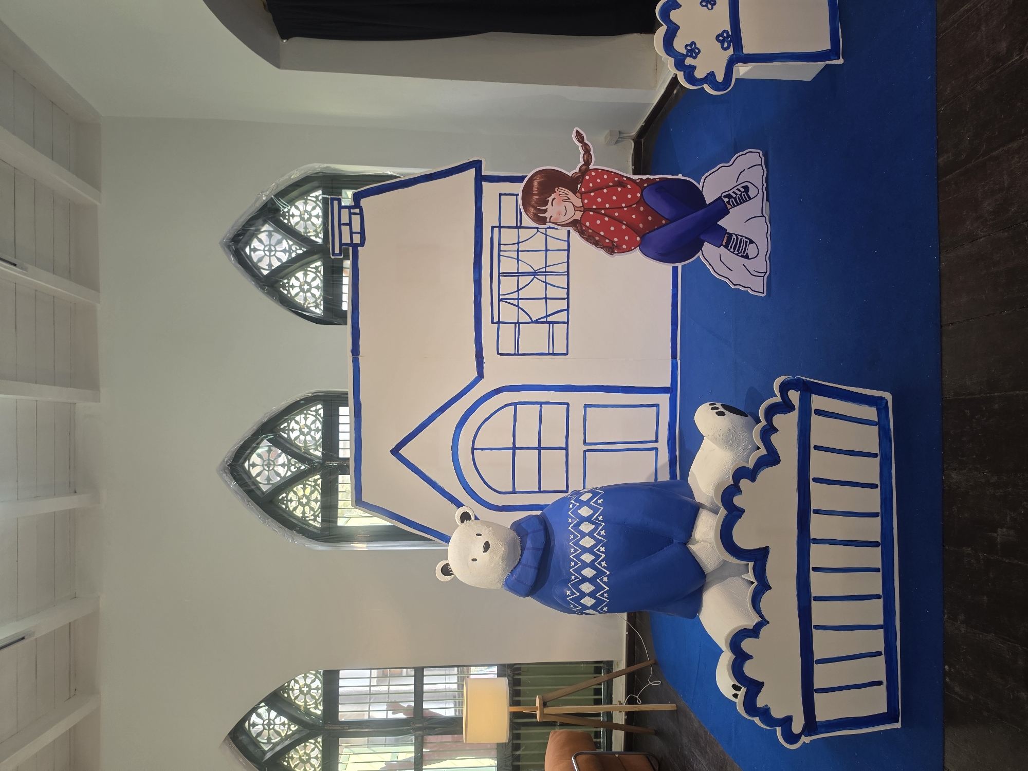 The scene is an indoor setup with a large blue floor covering. There are life-sized cardboard cutouts of a house, a girl, and a polar bear, all in a cartoonish style. The house is white with blue outlines, and the girl is wearing a red polka-dot shirt and blue pants. The polar bear is wearing a blue sweater with white designs. The backdrop features Gothic-style windows with intricate floral designs. A floor lamp is visible on the left side, along with a piece of furniture, potentially a chair, hinting at an interior design or display setting. The overall impression is that of a children's event or display. It's not possible to ascertain the location as Bangkok, Thailand from the content of the image.