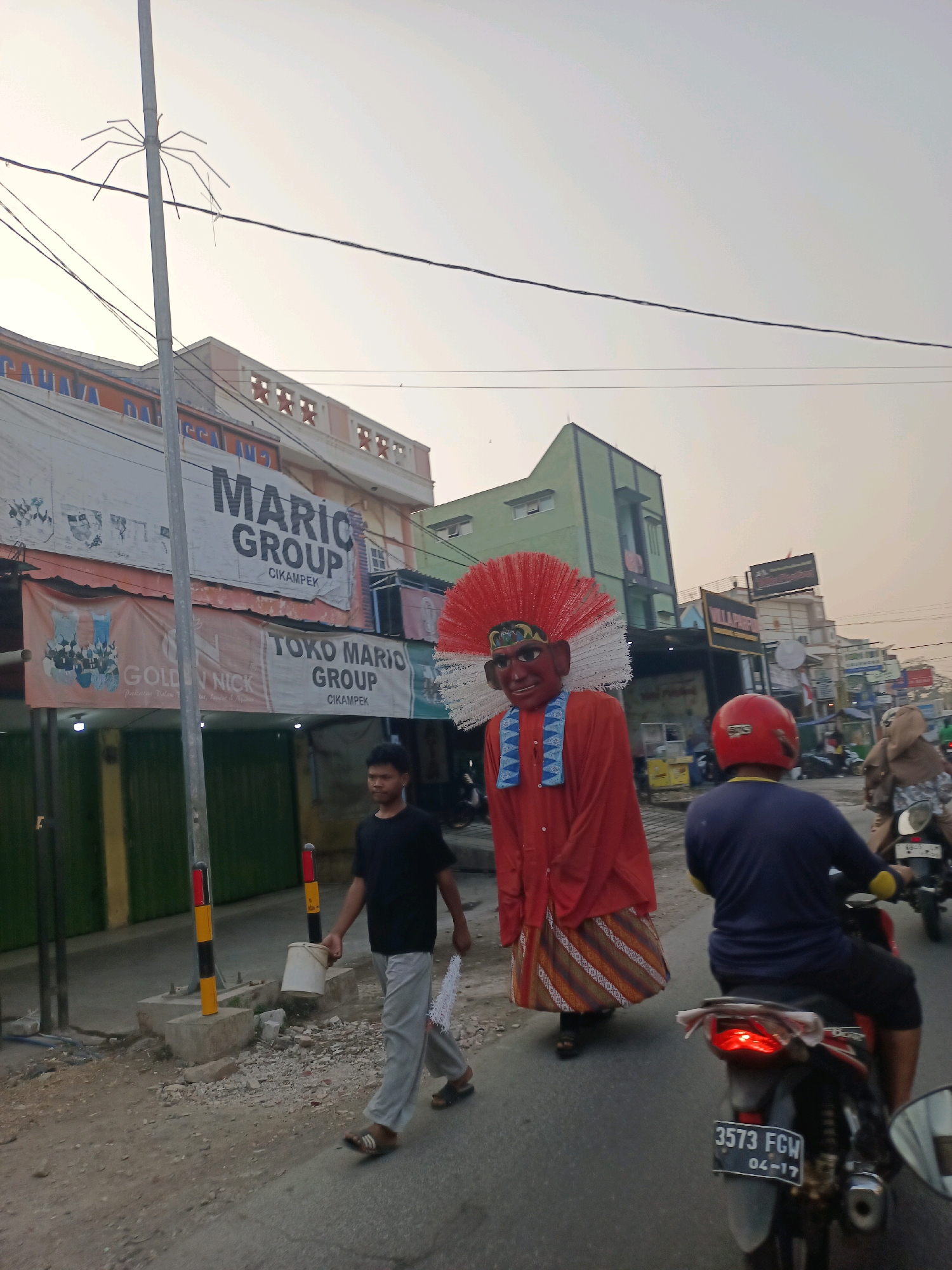 The image depicts a street scene in Cikampek, Karawang, Indonesia, during the day, likely early morning or late afternoon due to the soft, diffused light. In the foreground, a large, costumed effigy, likely an "Ondel-ondel" figure, is being moved along the street. The effigy is adorned with a prominent red feathered headdress, a red shirt, and a sarong with striped patterns. To the left of the effigy, a young man in a black t-shirt and grey pants walks carrying a white bucket and a white feathered object.
Several motorcycles are visible. One motorcycle in the mid-ground has a rider wearing a blue shirt and a red helmet, and another motorcycle behind it has a rider wearing a brown hijab. The street is paved with asphalt and appears to be a commercial area with various buildings lining the sides. Prominent signage includes "MARIO GROUP CIKAMPEK" and "TOKO MARIO GROUP CIKAMPEK". There are also banners and advertisements on the buildings. Electrical wires crisscross overhead, and a pole with a spider-like antenna is visible on the left. The environment appears somewhat dusty and unkempt, with debris on the side of the road.