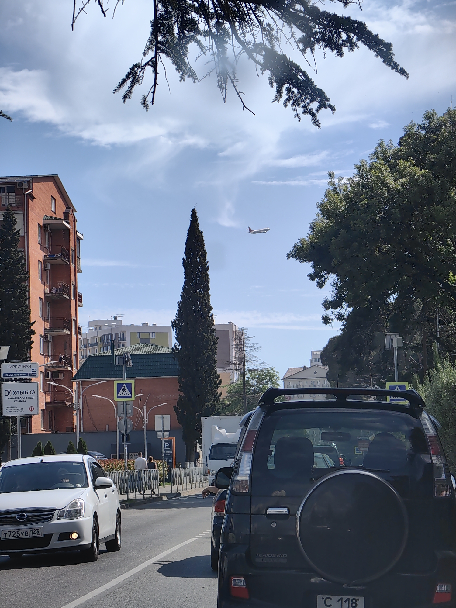 The image captures a sunny day in Sochi, Russia, with a clear blue sky and scattered clouds. A prominent feature is an airplane in flight, soaring high above the cityscape. In the foreground, a black SUV with a spare tire mounted on the back is stationary in traffic, its rear visible. To its left, a white Nissan sedan is also in traffic, its front visible. The backdrop includes several buildings, ranging from a red brick apartment complex with balconies to modern white structures, indicating an urban environment. Lush green trees, including a tall, slender cypress, line the street and frame the scene. Road signs are visible, suggesting a city street with traffic regulations. The overall impression is of a typical urban scene in Sochi, with a blend of residential buildings, greenery, and ongoing traffic.