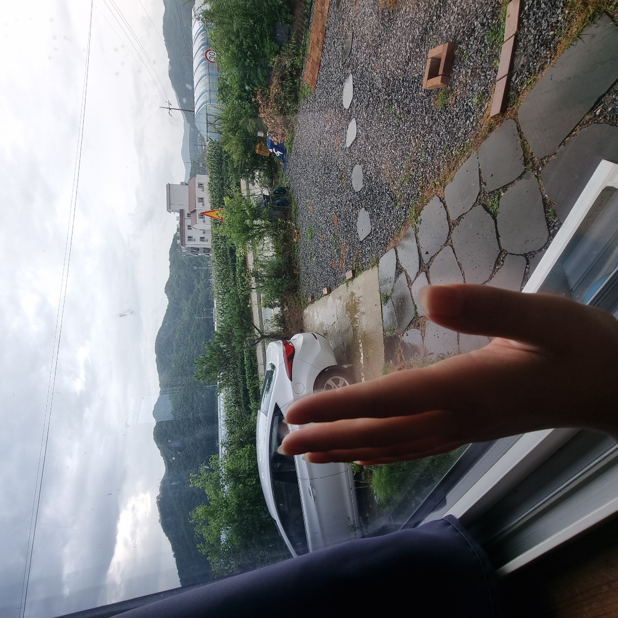A human hand is held up in the foreground, obscuring part of the view from a window. Through the window, a white car is parked on a wet concrete surface. The car is facing away from the viewer and appears to be a sedan. Beyond the car, a path made of large, flat stones is visible, leading into a gravel-covered area. The background consists of a lush green landscape with trees and rolling hills under a cloudy sky. In the distance, there are some buildings, including what appears to be a greenhouse with a "30" speed limit sign. The scene is outdoors, likely in a rural or semi-rural setting. The overall impression is of a slightly overcast or rainy day. The location is identified as Dunnae-myeon, South Korea.