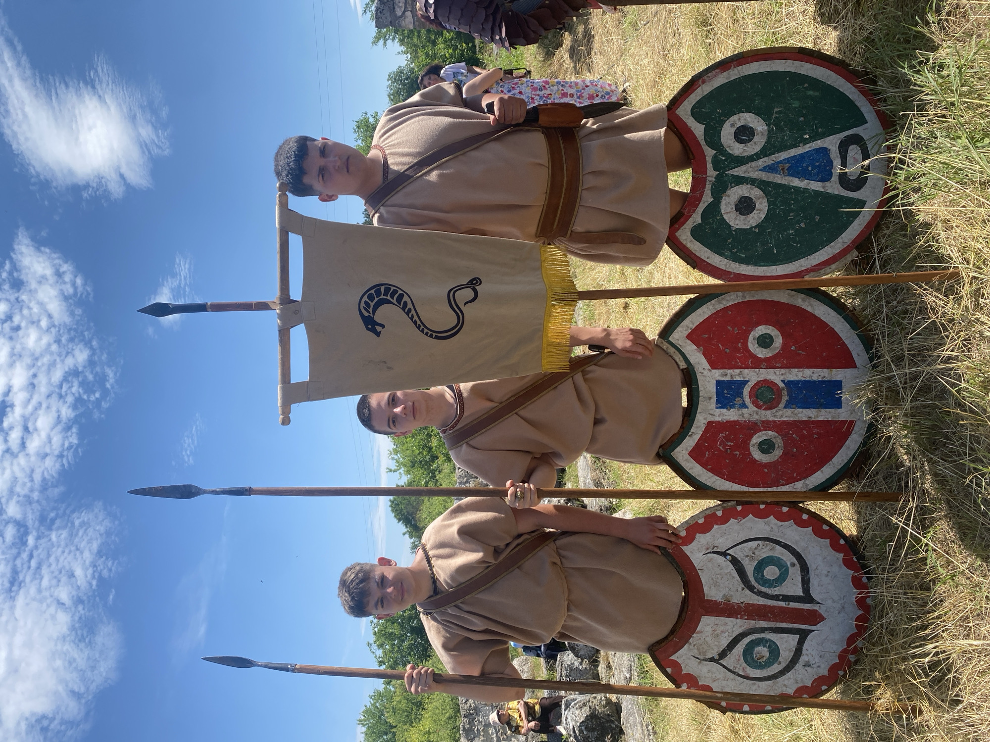 Three young males, appearing as teenagers or young adults, are depicted standing outdoors in what appears to be an archaeological site in Gigen, Bulgaria, under a clear blue sky with scattered white clouds. All three are dressed in historical-style costumes consisting of light brown, knee-length tunics with short sleeves and dark brown baldrics (shoulder straps).
From left to right:
1. **Leftmost Individual:** Poses with a slight smile, holding a tall wooden spear with a dark metal tip in his right hand. A circular shield, predominantly white with a red and black stylized mask or owl-like face design, rests on the dry grass in front of him.
2. **Middle Individual:** Stands with a neutral expression, holding a similar wooden spear with a metal tip in his right hand. He wears a prominent gold-colored ring on the ring finger of his left hand. A circular shield, featuring a red and white background with a central blue vertical stripe and a red circle, rests at his feet.
3. **Rightmost Individual:** Poses with a serious expression. He holds a light beige banner on a wooden pole in his right hand. The banner displays a black stylized coiled snake or serpent design and has a yellow fringe along its bottom edge. He also wears a dark brown belt and holds a sheathed knife or dagger in his left hand, which is attached to his belt. A circular shield, predominantly dark green with a white and blue stylized face design, rests on the ground.
The ground is covered with dry, light green and yellow grass. In the background, out-of-focus elements include ancient-looking stone ruins or large rocks, green foliage from trees and bushes, and faint hints of distant structures or utility lines. Several other blurred individuals are partially visible in the mid-ground, some appearing to be seated on stones, suggesting a public event or gathering at the site. The overall impression is a historical reenactment or exhibition.