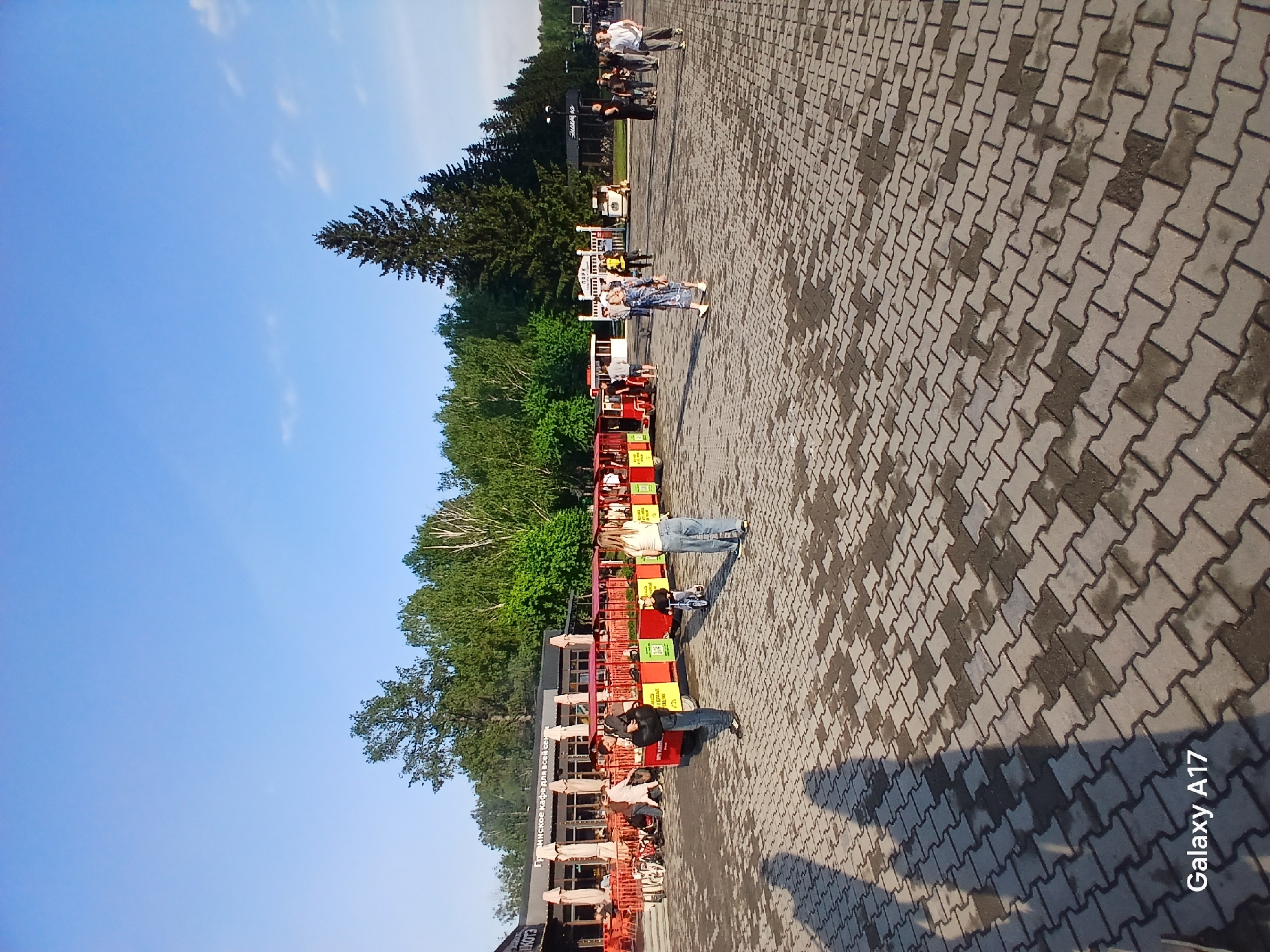 The image depicts a scene in Yekaterinburg, Russia. A paved area, possibly a public square or plaza, shows patterns of wetness or dampness. In the background, there's a line of trees and what appears to be an outdoor restaurant or cafe with orange fencing and multiple people seated. Individuals are also walking around the paved area. The sky is visible with a clear blue tone. A train is visible in the shot with people sitting inside. The lower right corner shows the text "Galaxy A17".