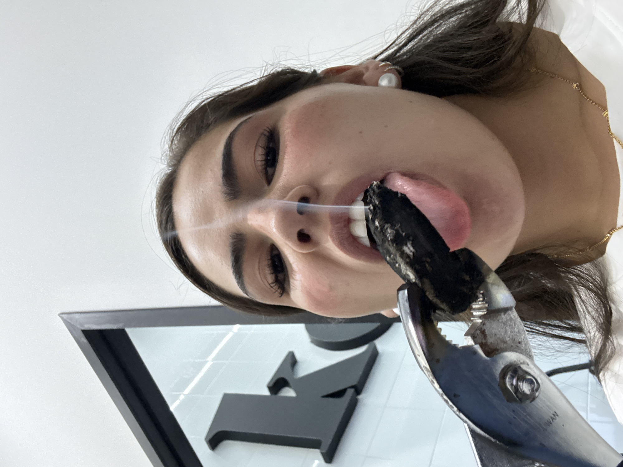 A woman with dark hair and fair skin is looking directly at the camera with an unusual perspective, as if the camera is positioned below her. She is holding a dark, smoking object in her mouth, held by pliers. She is wearing a pearl earring and a gold necklace. The background appears to be a white tiled wall with a black logo. The specific location in San Bernardino Tlaxcalancingo, Mexico cannot be determined from the image.