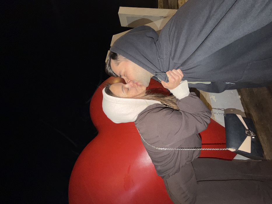 The image displays a nighttime scene in Tel-Aviv, Israel, specifically at the "I ♥ TLV" public art installation. Two individuals, a female and a male, are engaged in a kiss.
The female, positioned on the left, is leaning forward towards the male. She wears a brown hooded sweatshirt with a light-colored interior hood lining, and dark pants. A black and light beige cross-body purse with a silver chain strap is visible at her side. Her eyes are closed, and her lips are pressed against the male's.
The male, positioned on the right, wears a dark hooded zippered sweatshirt with the hood pulled up. His eyes are closed, and his lips are pressed against the female's. His left hand (viewer's perspective) holds the female's right hand, which is gripping the fabric of his sweatshirt near the zipper. A ring is visible on his left ring finger.
Behind the female, a large, illuminated red heart sculpture, part of the "I ♥ TLV" sign, occupies the left background. To the right, behind the male, the letters "L" and "V" of the "TLV" portion of the sign are visible, appearing as lighter-colored, block-like structures. The upper background is dark, indicating a nighttime setting.