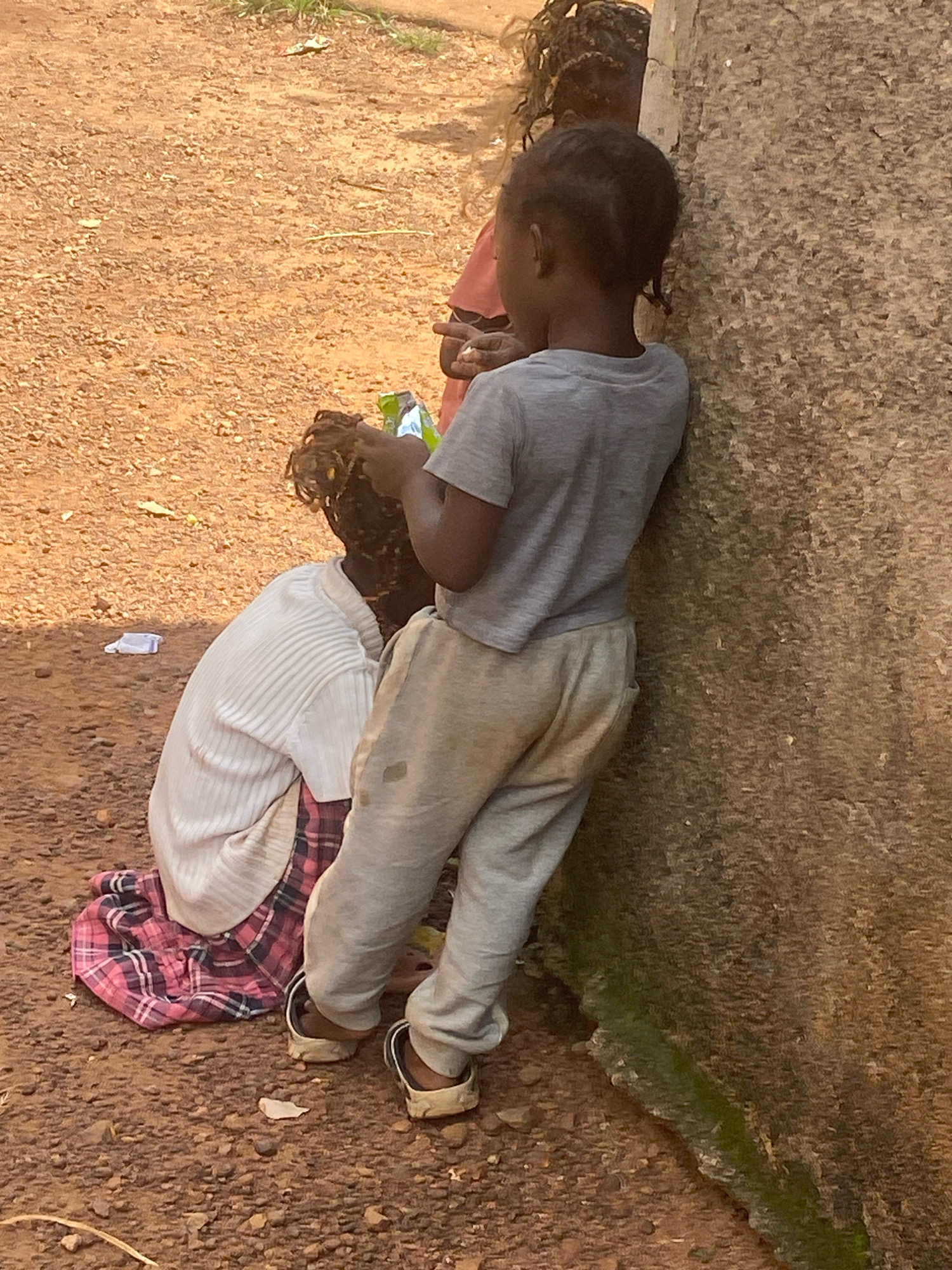 In Bukuru, Nigeria, two children are near a textured concrete wall. One child, wearing a grey t-shirt and light-colored pants, leans against the wall, facing away from the camera. The other child, seated on the ground and facing the wall, wears a white ribbed long-sleeved shirt over a pink and blue plaid skirt. The ground is a rough, reddish-brown dirt surface with scattered small stones and a white paper object. The wall appears weathered, with patches of green moss at its base. Another child's back is visible in the background, partially obscured by the child leaning against the wall.
