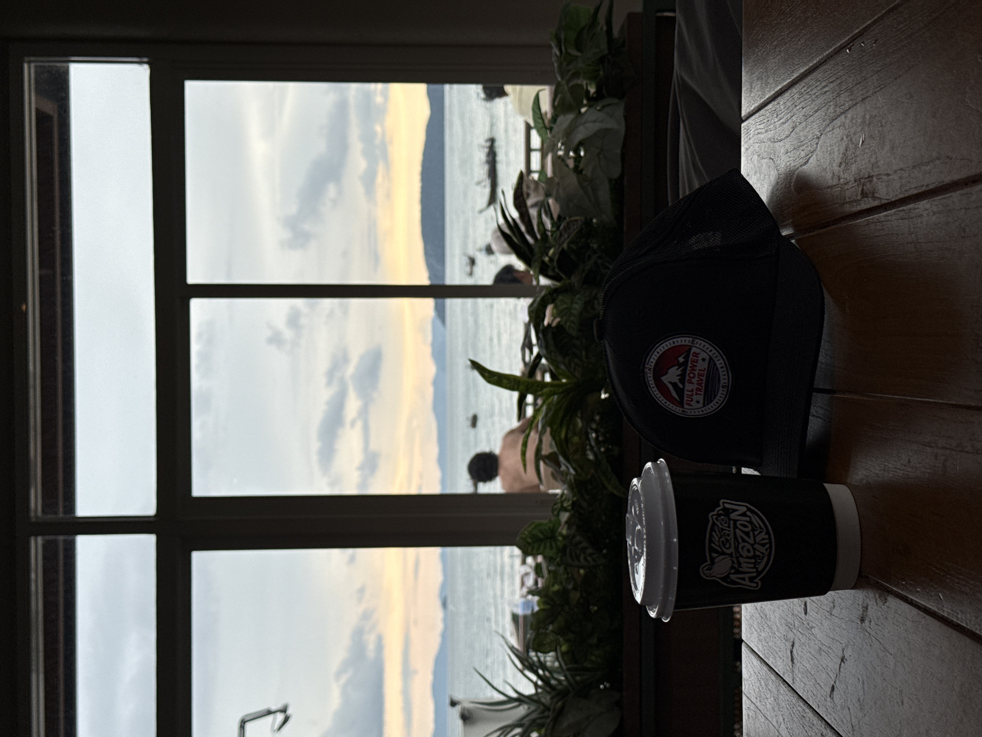 A black coffee cup with a white lid sits on a wooden table next to a black cap with a circular emblem. Behind the table, through a window with dark frames, a body of water is visible under a cloudy sky. The sky transitions to yellow and orange hues near the horizon, suggesting either dawn or dusk. Several islands or landmasses are visible in the distance. The water is dotted with a few small boats, some appearing to be long-tail boats, a common sight in Thailand. In the middle ground, there are several people seated, their backs to the camera, facing the water. Lush green plants are arranged in a planter box in front of the window. The overall impression is a relaxed atmosphere with a view of the ocean. The presence of Cafe Amazon branding on the cup suggests the location is a Cafe Amazon, a popular coffee chain in Thailand, likely in or near Ban Ao Nang, Krabi province, which is known for its scenic coastal views and islands.