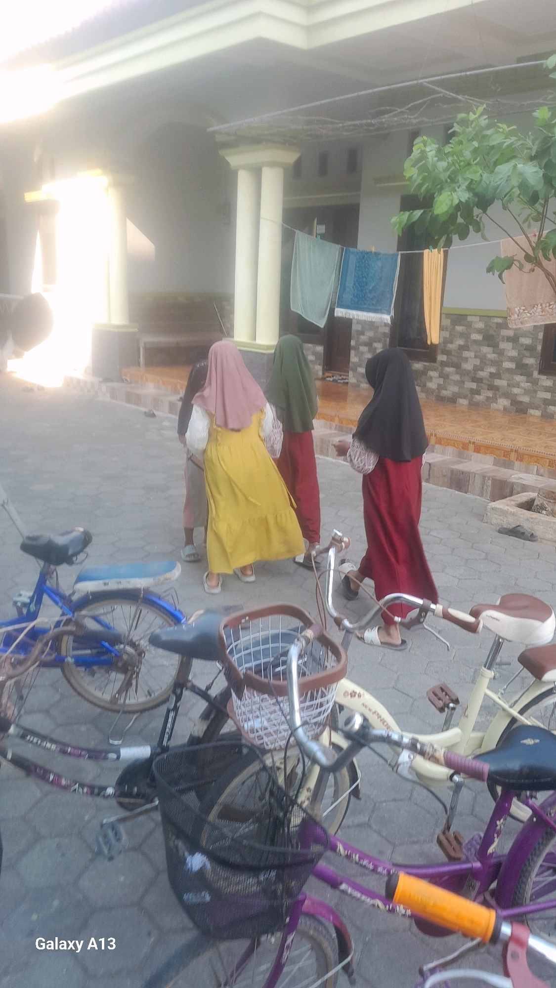 The image depicts a group of three young women standing in front of a building, likely a house, in Ngelo, Indonesia. The women are all wearing hijabs and modest clothing. One woman on the left is wearing a pink hijab and a yellow dress. The woman in the middle is wearing a green hijab and a dark red outfit. The woman on the right is wearing a black hijab and a dark red skirt. They appear to be gathered and possibly conversing. In the foreground, there are several bicycles parked, some with baskets. A blue bicycle is to the left, and a light-colored bicycle with a brown basket is to the right, with a purple bicycle partially visible at the bottom. The environment suggests an outdoor residential setting, with a paved ground and part of a building with pillars and windows visible in the background. Laundry is hanging on a line in front of the building. The lighting indicates it is daytime, possibly late afternoon, with sunlight casting a warm glow.