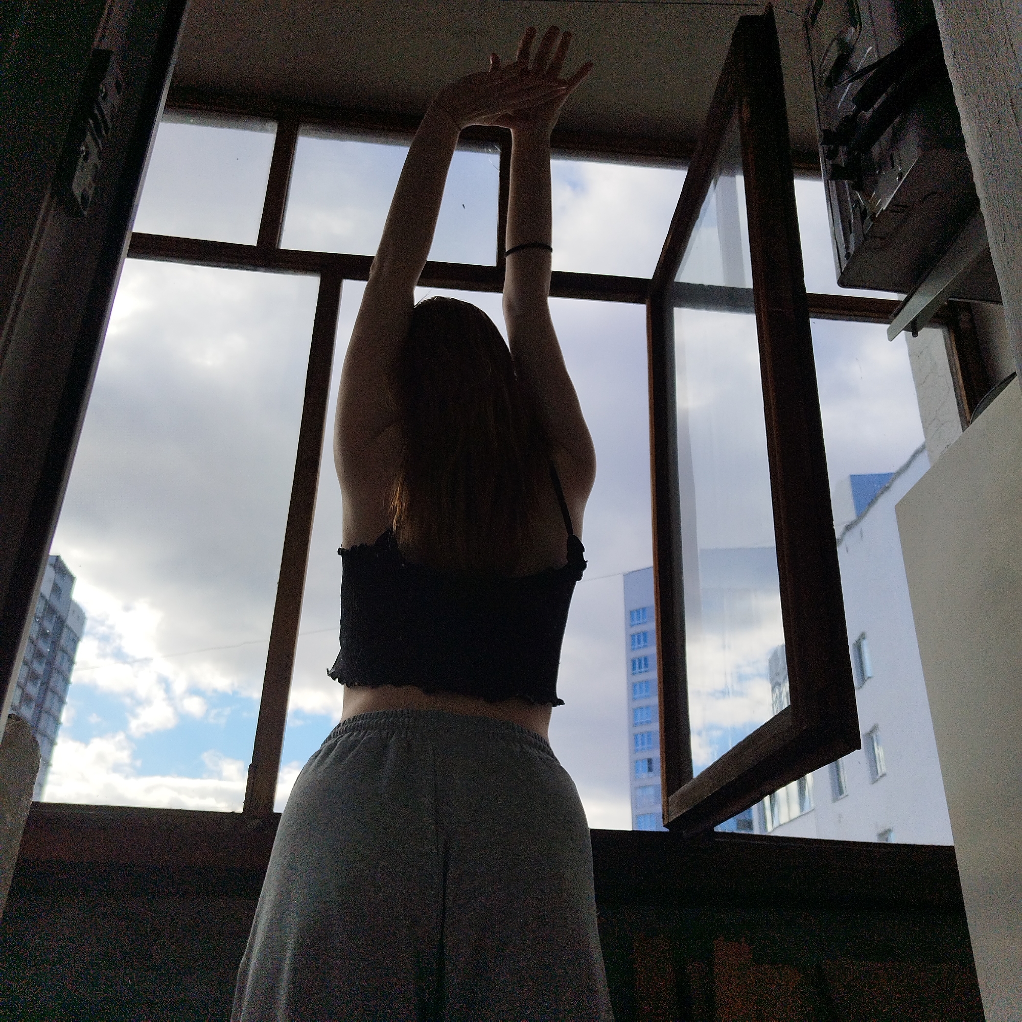 The image depicts a woman, seen from the back, stretching her arms upwards in front of an open window. She is wearing a dark crop top and grey sweatpants. The window frames a view of the city of Yekaterinburg, Russia, characterized by high-rise buildings with a cloudy sky above. An open window frame is to the right.