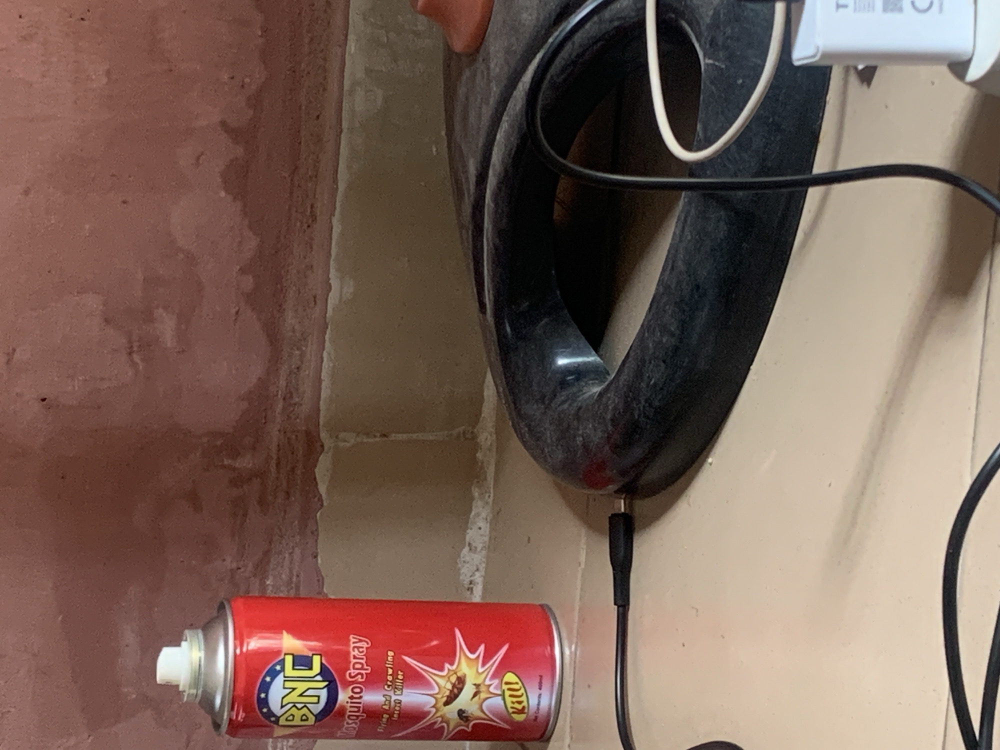 The image depicts a close-up view of a can of "BNC Mosquito Spray" placed on a tiled surface. The can is predominantly red with a white cap and features a circular logo with the letters "BNC" in yellow and blue. Text below the logo reads "Mosquito Spray," "Flying And Crawling Insect Killer," and "Kill!". Below this, it indicates "Net Contents: 400ml". To the right of the can, a black, circular object, possibly part of a device or appliance, is visible. A black cable extends from the left of the frame towards this object, with a connector plugged in. Another cable, white in color, curves from the right side of the black object and disappears out of frame. A white power adapter with some lettering and symbols is also visible in the lower right corner. The background features a textured, reddish-brown wall. The overall scene appears to be indoors, likely in a residential setting. There is no indication of the city of Jauro Yinu, Nigeria, in the image itself.