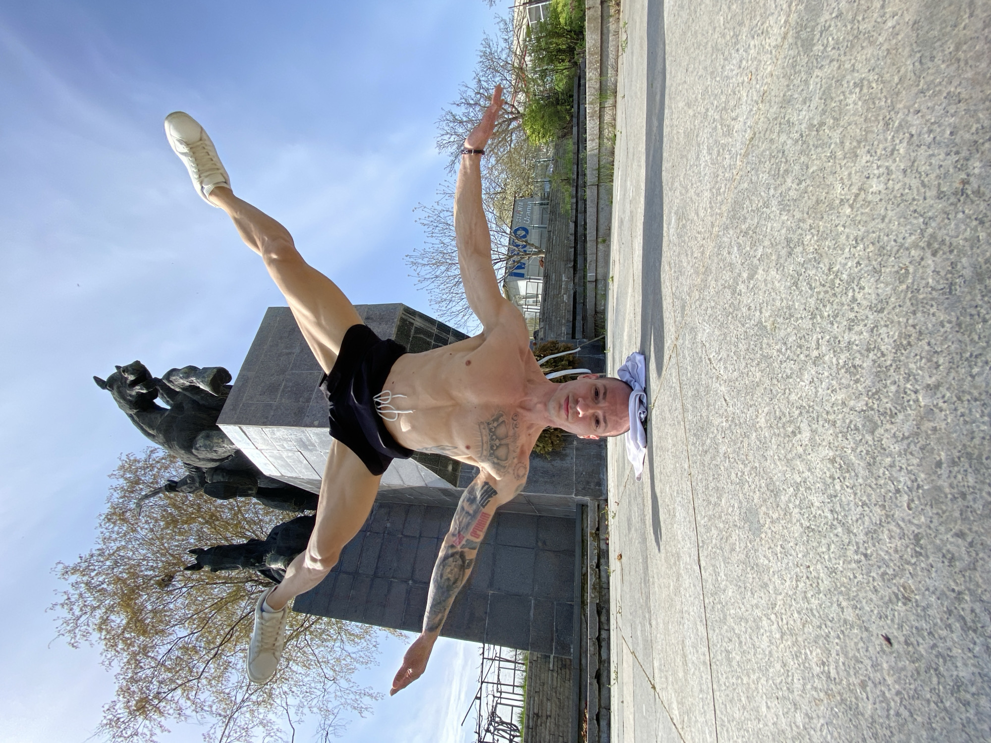 A man is photographed at ground level in an outdoor scene. He is balancing on his head, with his legs extended outwards at roughly a 90-degree angle. He is wearing black shorts and white sneakers. His arms are outstretched, with one pointing upwards and the other downwards. The man has several tattoos on his arms and chest. His head is resting on a white towel placed on a grey paved area.
To the left of the man is a monument depicting horses, possibly the Horsemen Monument in Pleven. Behind him, buildings and trees are visible under a clear blue sky.
