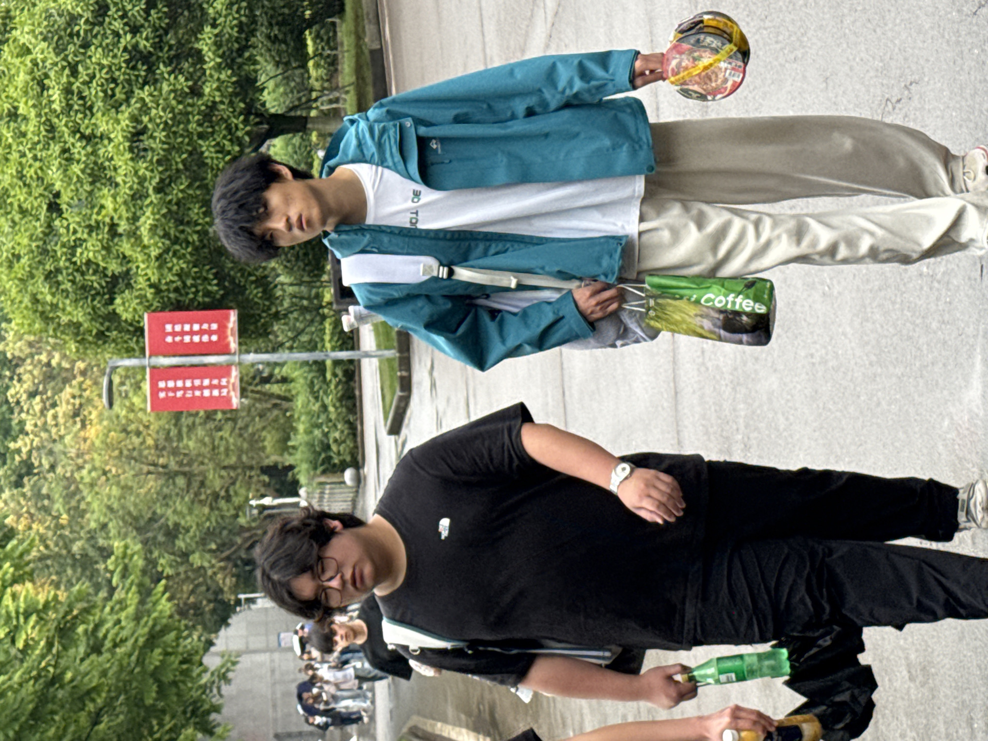 The image shows two young men on a city street in Jiangjin, China. The first man, positioned at the top of the frame, wears a teal jacket over a white t-shirt and light-colored pants. He carries a container of food in one hand and a bag labeled "coffee" in the other. The second man, at the bottom of the frame, wears a black t-shirt and black pants, with glasses and a watch on his left wrist. He is holding several bottles. They are in front of a large green area. A red sign with illegible writing is in the background. Several other individuals are walking in the background.