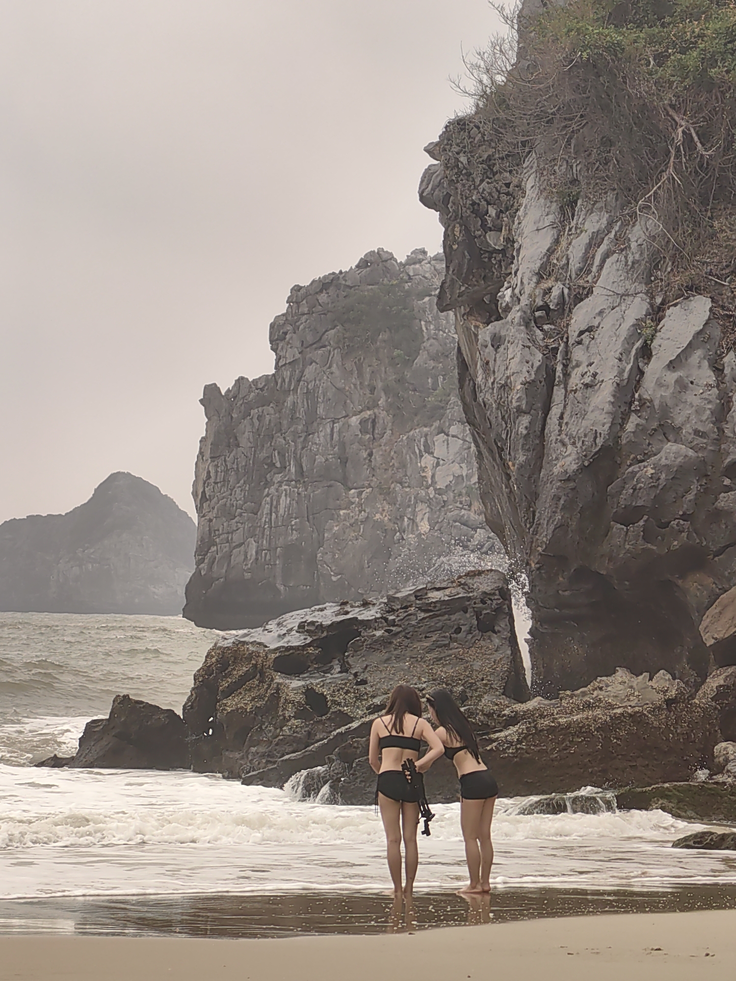 Two women in black bikinis stand on a sandy beach at Cát Bà Island in Hải Phòng, Vietnam. They face rocky cliffs where waves crash against the shore. The sea is choppy under an overcast sky with rocky islands visible in the distance. The women appear to be observing the water cascading from a crack in the cliff face. One woman holds what appears to be a pair of flippers or water shoes. The terrain is characterized by jagged rock formations and sparse vegetation.