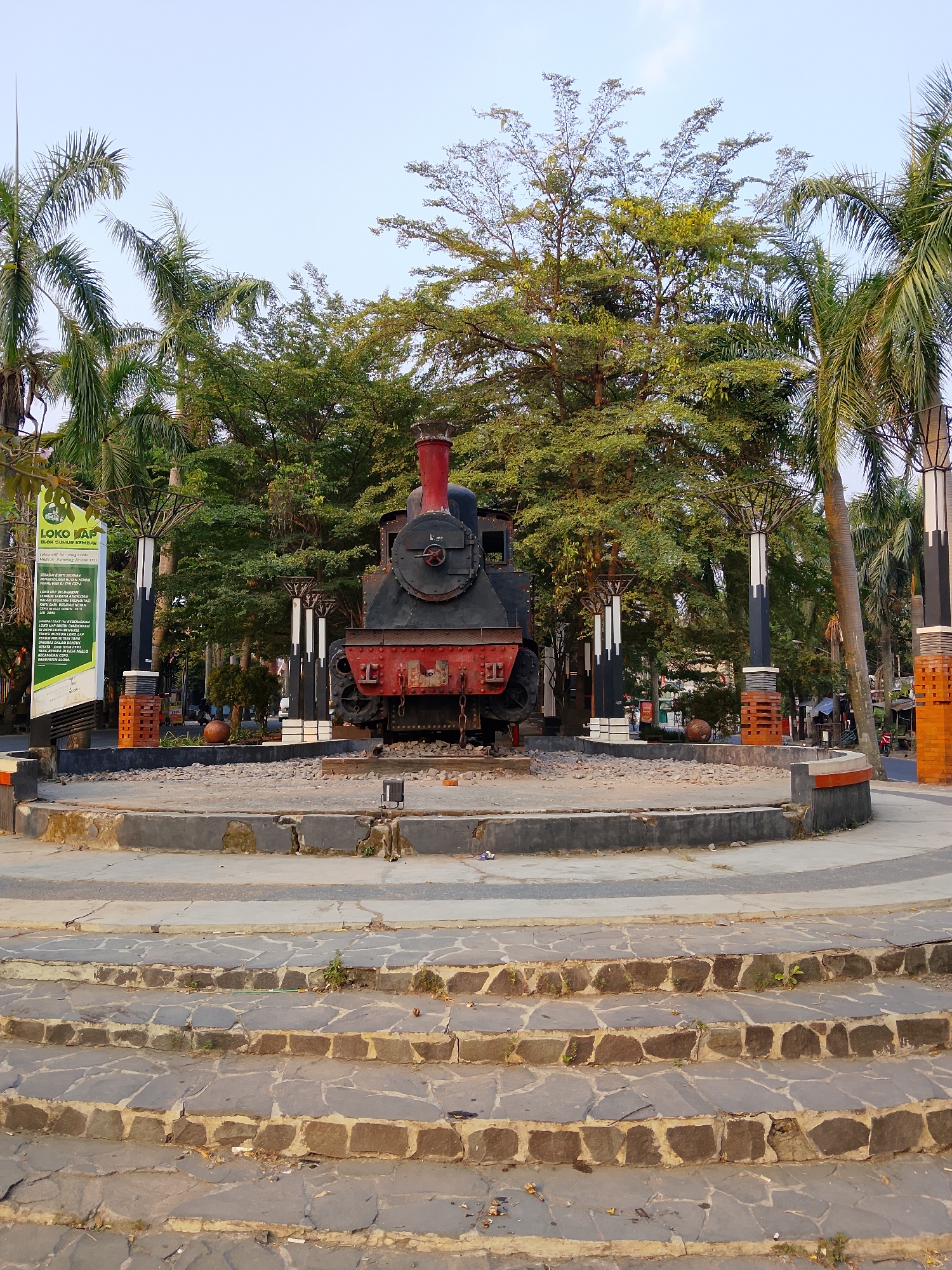 A vintage steam locomotive is on display in a circular plaza at the Loko Via heritage site in Balun, Indonesia. The locomotive is black with red accents and a tall smokestack. The plaza is surrounded by a low concrete wall and is paved with gravel. Steps made of irregular stone blocks lead up to the plaza from the foreground. In the background, palm trees and other foliage create a lush green environment. Several decorative pillars, tall and slender with white and black sections, are placed around the plaza. A green sign on the left side of the image provides information about the site. The sky is clear and pale blue.