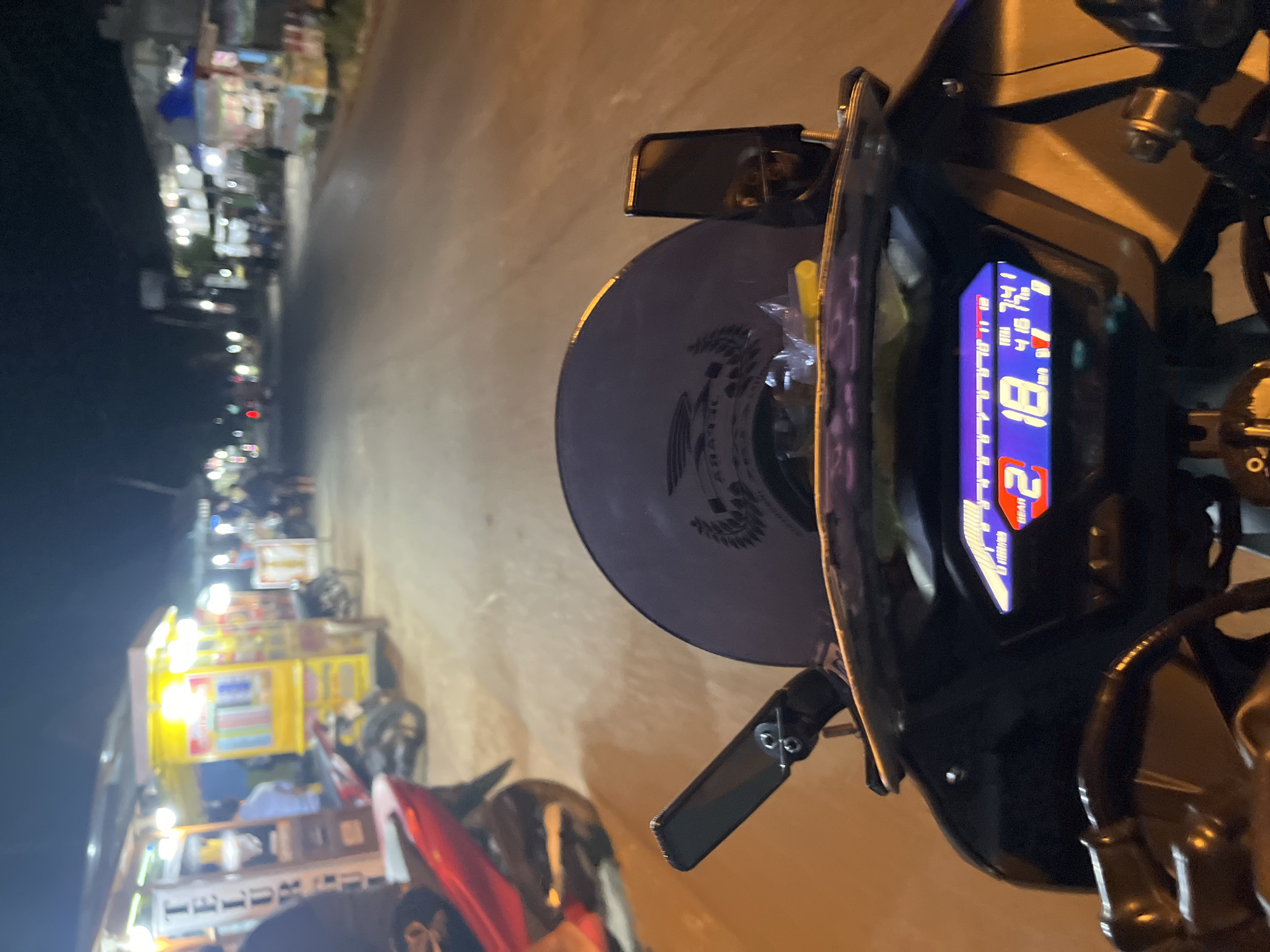 The image is a first-person perspective from a motorcycle at night in Pecangaan, Indonesia. The motorcycle's digital dashboard is visible in the foreground, displaying "2 18." and other data. To the left, a red motorcycle is parked. The background shows a street lined with food stalls and shops, illuminated by artificial lights. Several people are visible near the stalls, and a few other motorcycles are parked or in motion on the street. The overall scene depicts a lively night market or street food area.